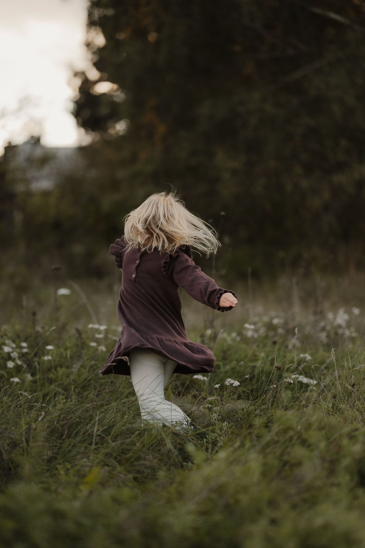 A person in a brown sweater and white boots spins joyfully in a grassy meadow at dusk.