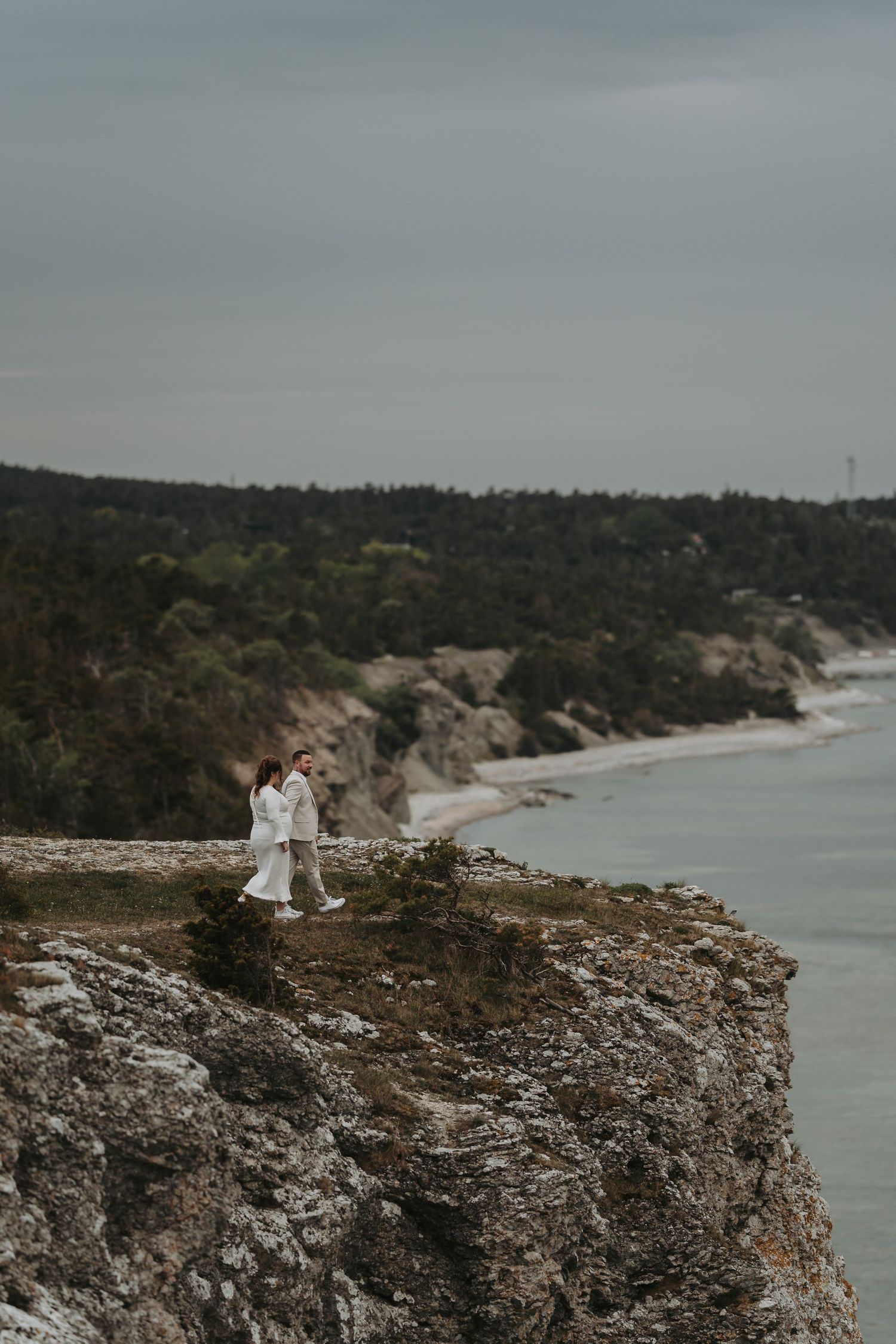 Two people stand together on a dramatic coastal cliff overlooking a scenic bay with forested shoreline.