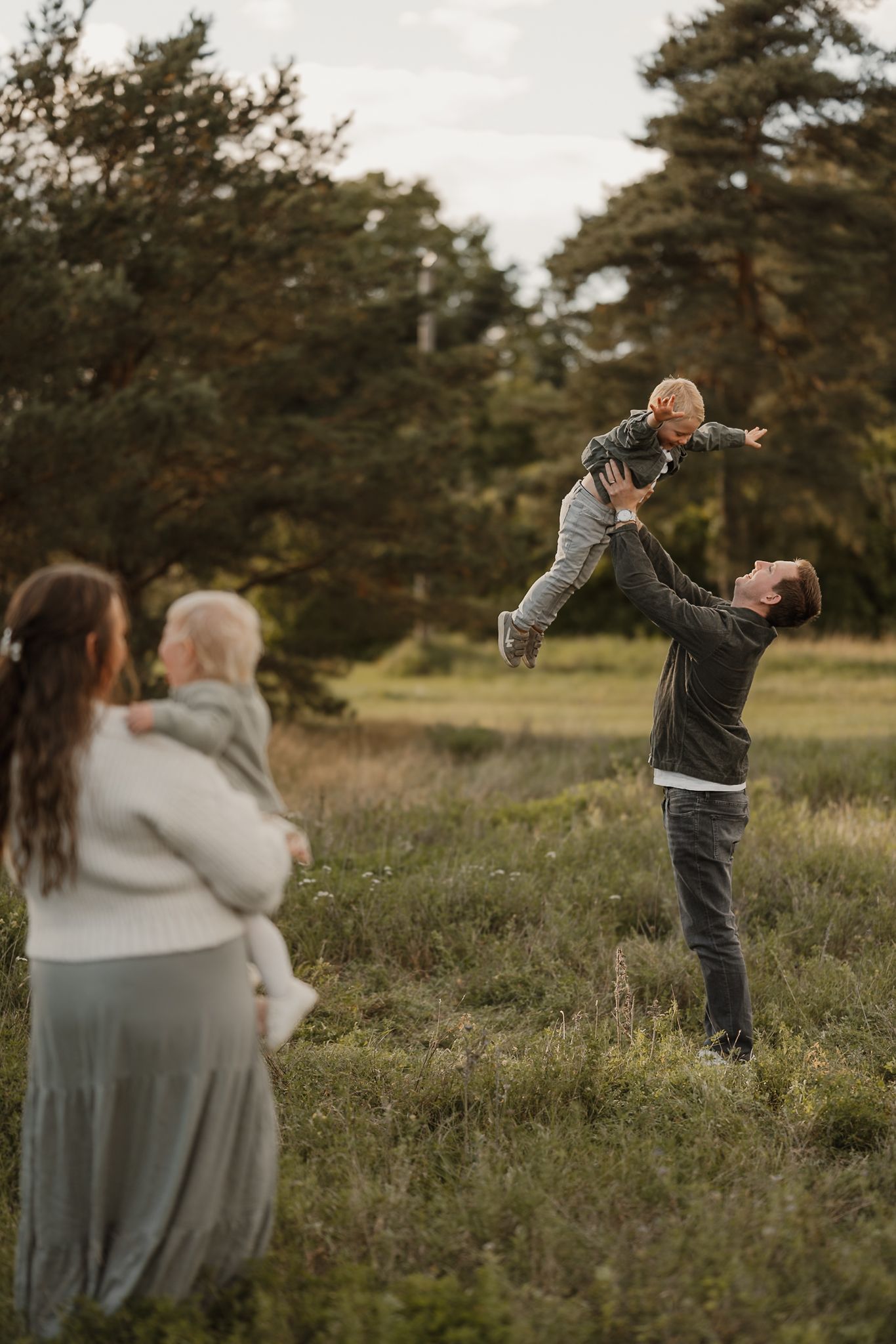 A father joyfully tosses his toddler in the air outdoors while a mother holds another child in a lush green field.