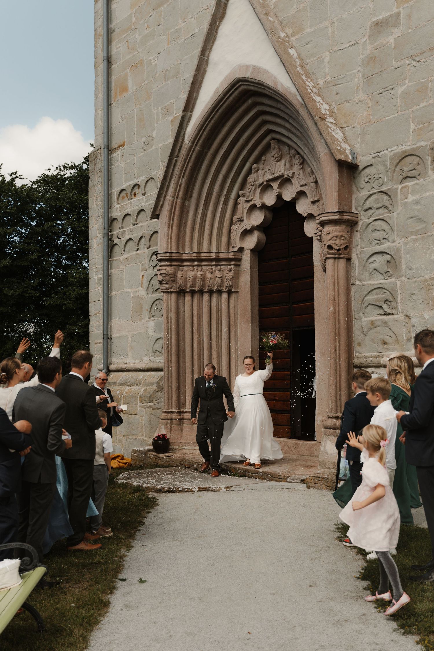 Newlywed couple exits a Gothic stone church doorway, greeted by cheering guests lining the path after their wedding ceremony.