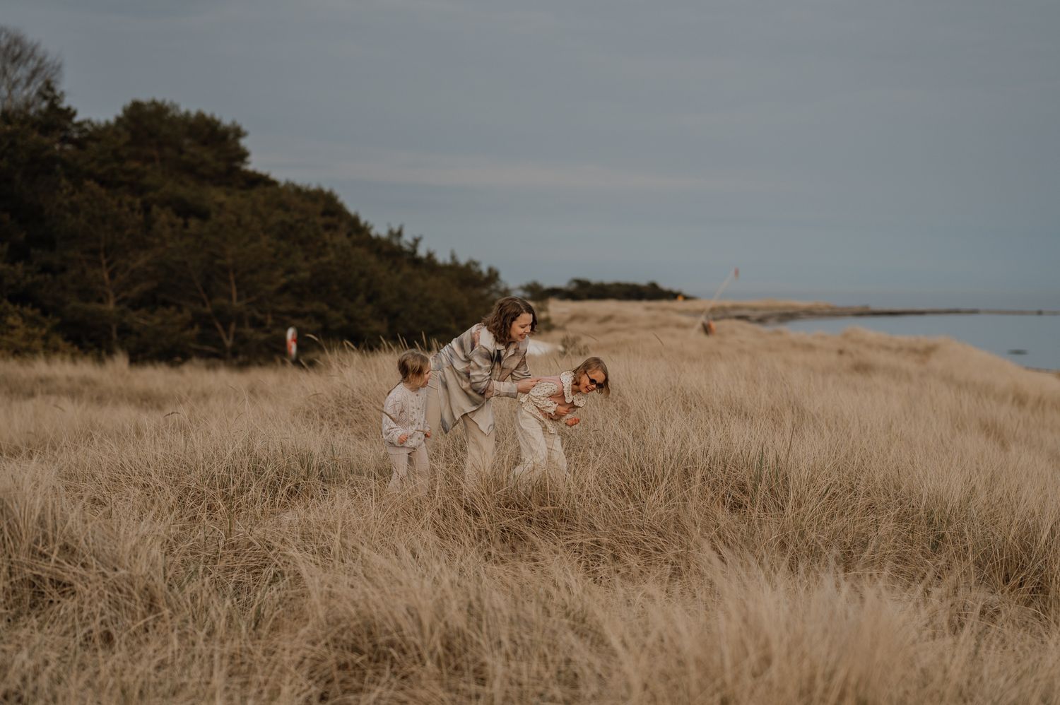 A mother and two children walk through golden beach grass near a coastal shoreline on a cloudy day.