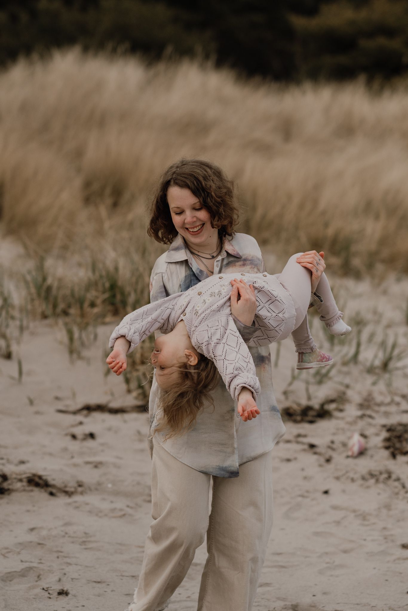 A laughing woman playfully holds a toddler upside down on a sandy beach with golden grass in the background.
