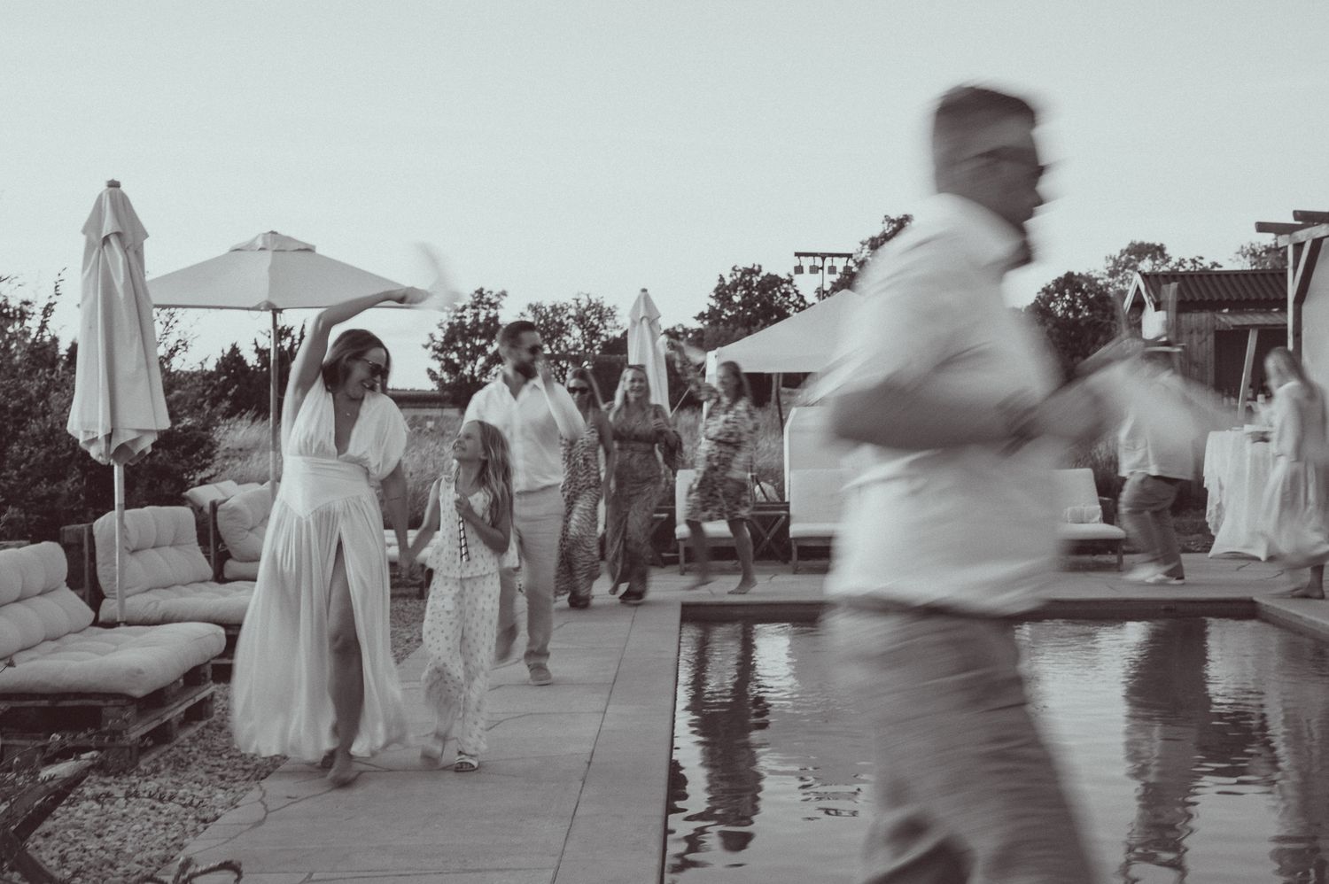 Black and white outdoor wedding reception guests mingling near a pool with patio umbrellas in background.