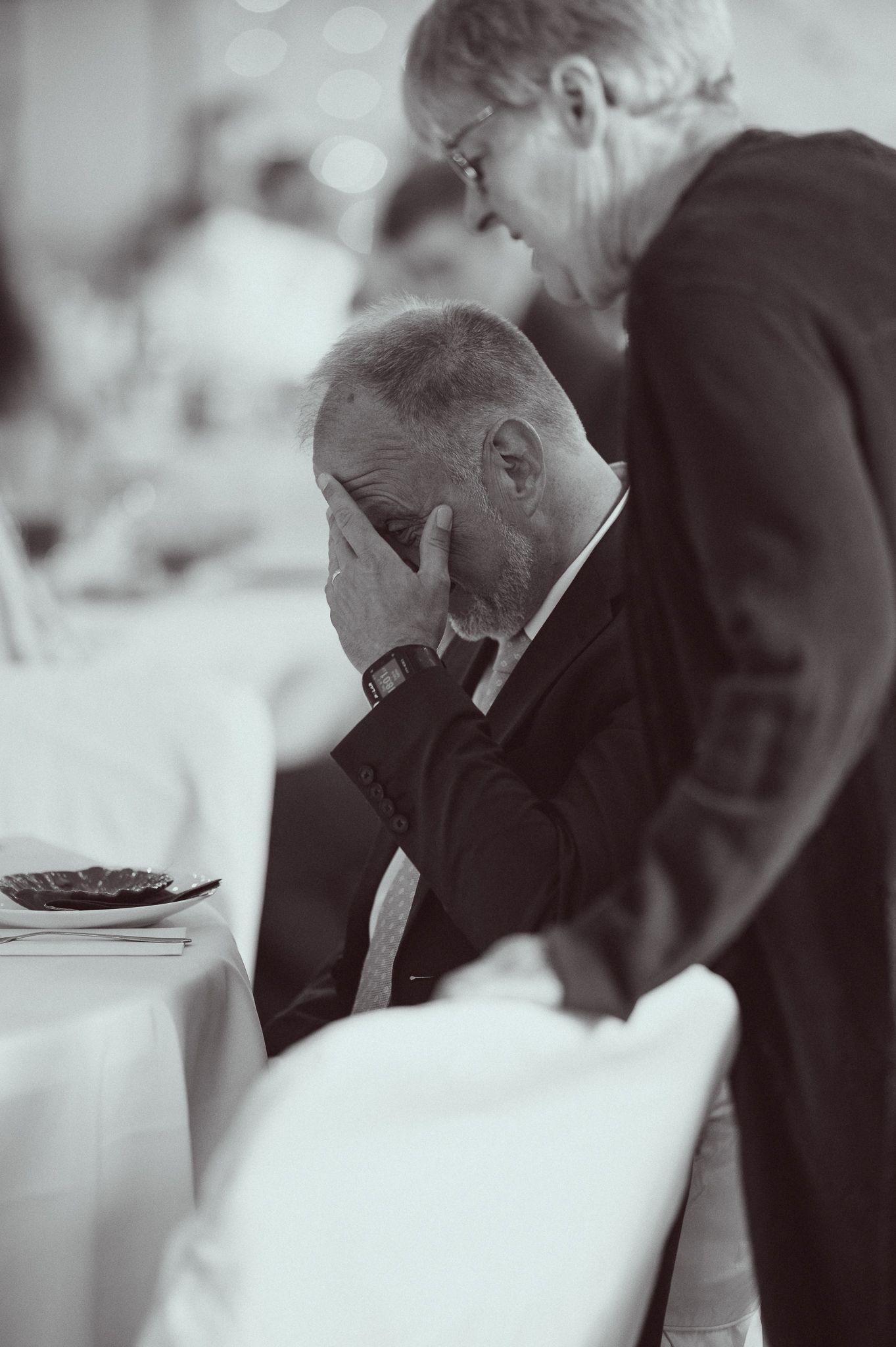 Black and white photo of an emotional man covering his face at a formal event, with a woman standing beside him.