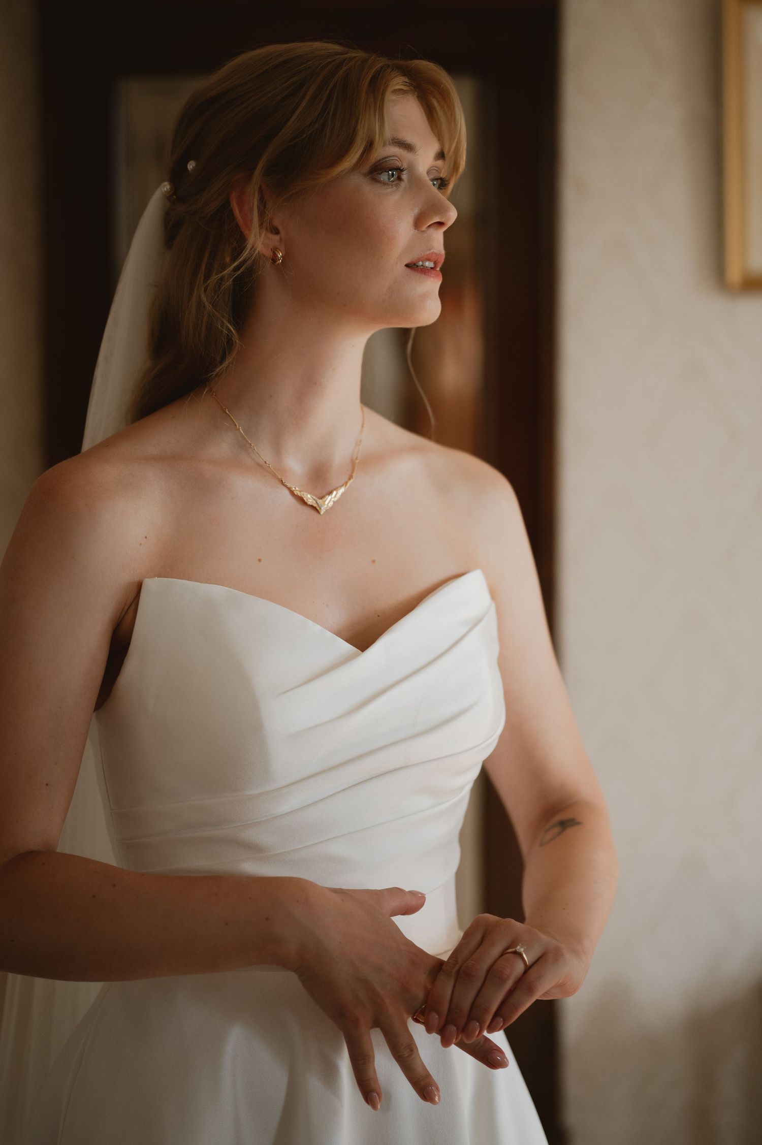 Elegant bride in strapless white wedding gown with veil and gold necklace gazing thoughtfully toward natural window light.
