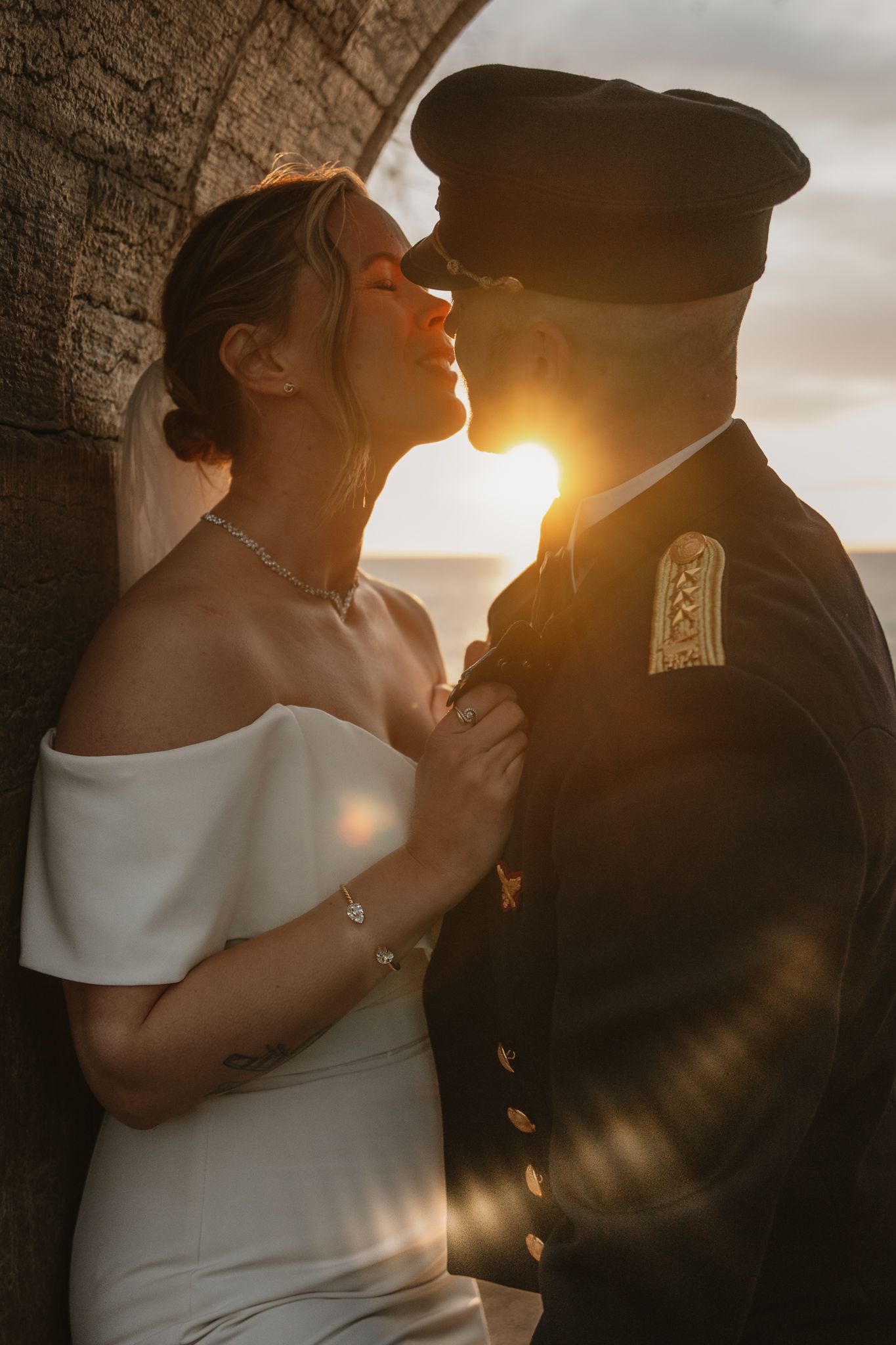 Bride and groom share a romantic sunset kiss, she in white off-shoulder gown, he in military dress uniform.