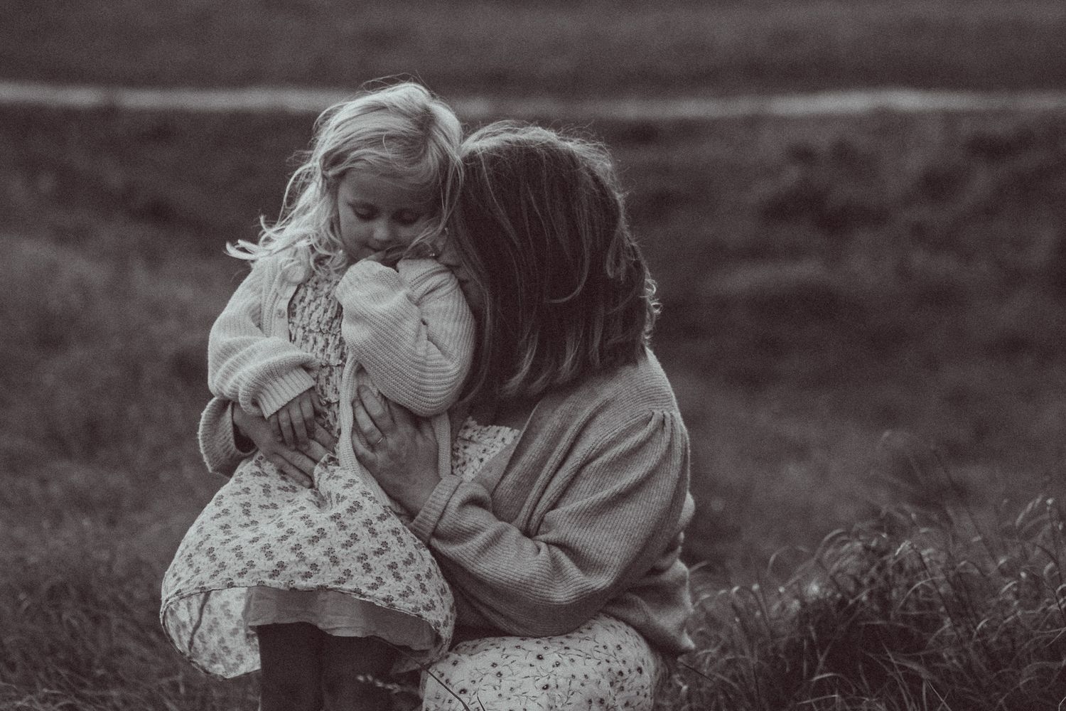 Black and white photo of a mother tenderly holding and kissing her young blonde toddler daughter outdoors.