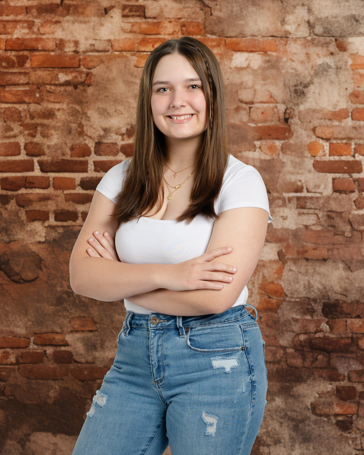 A person wearing a white top and distressed jeans poses confidently against a rustic brick wall background.