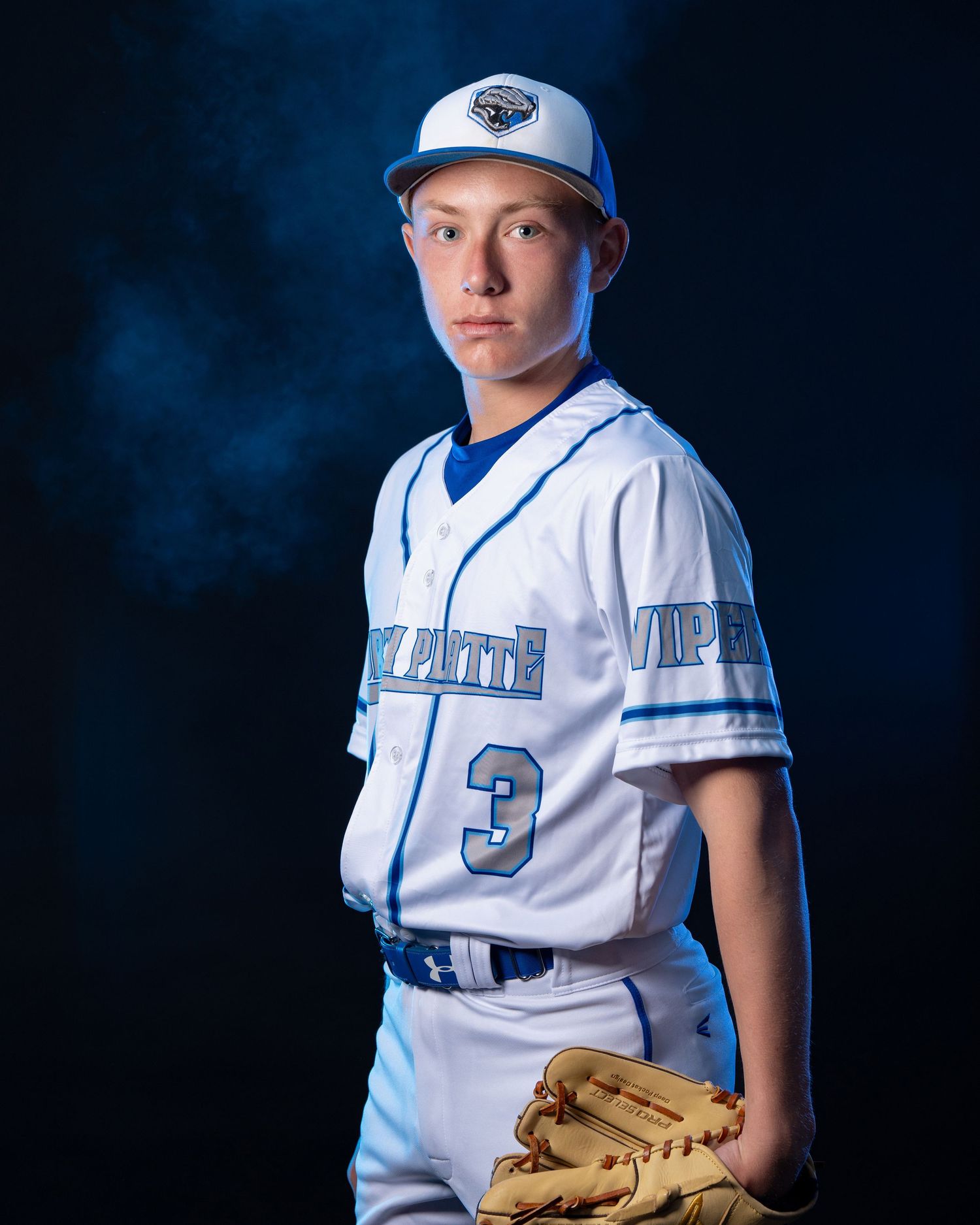 Baseball player in white and blue uniform poses with glove in dramatic studio lighting against dark background.