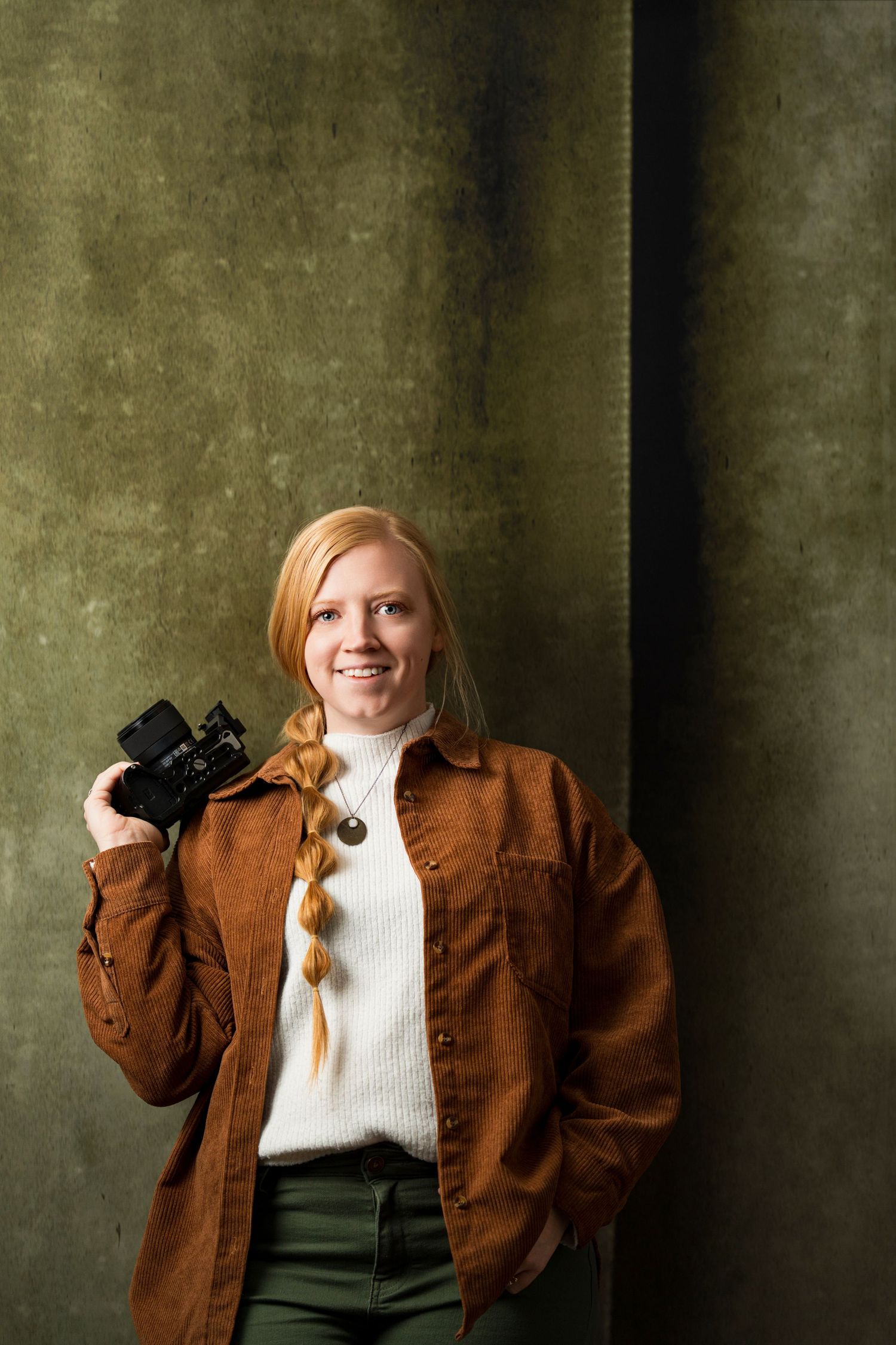 A person in a brown jacket and white sweater poses against a textured wall in a photo series.