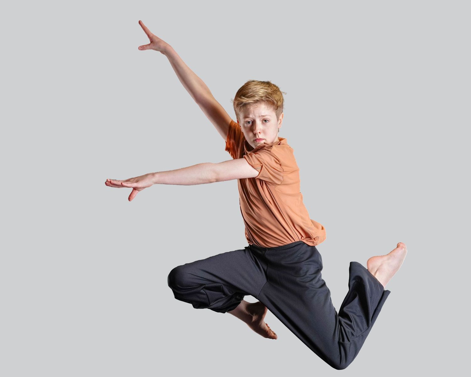 Young dancer in orange shirt and navy pants leaps gracefully against white background with arms outstretched.