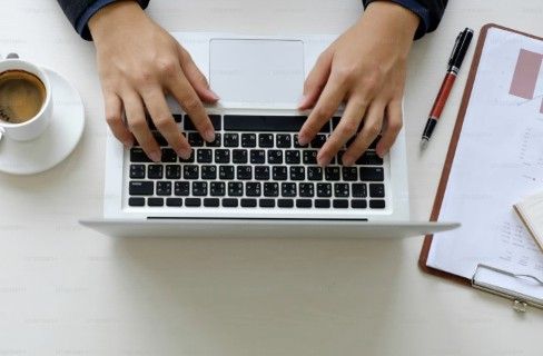 Overhead view of hands typing on a laptop with coffee cup, notebook, and pen on a white desk.