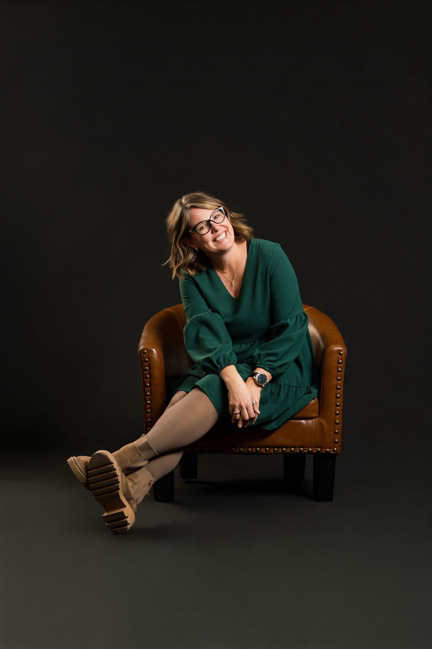 Professional studio portraits on a dark background featuring a brown leather chair and emerald green outfit styling.