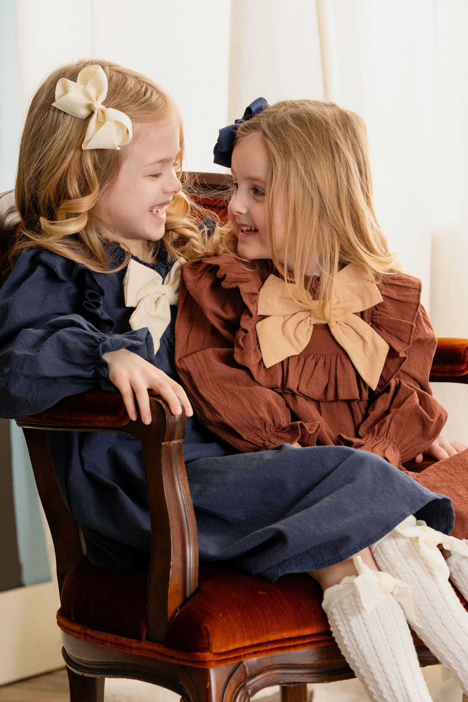 Two young children in vintage-style dresses with bows share a sweet moment while sitting on a wooden chair.