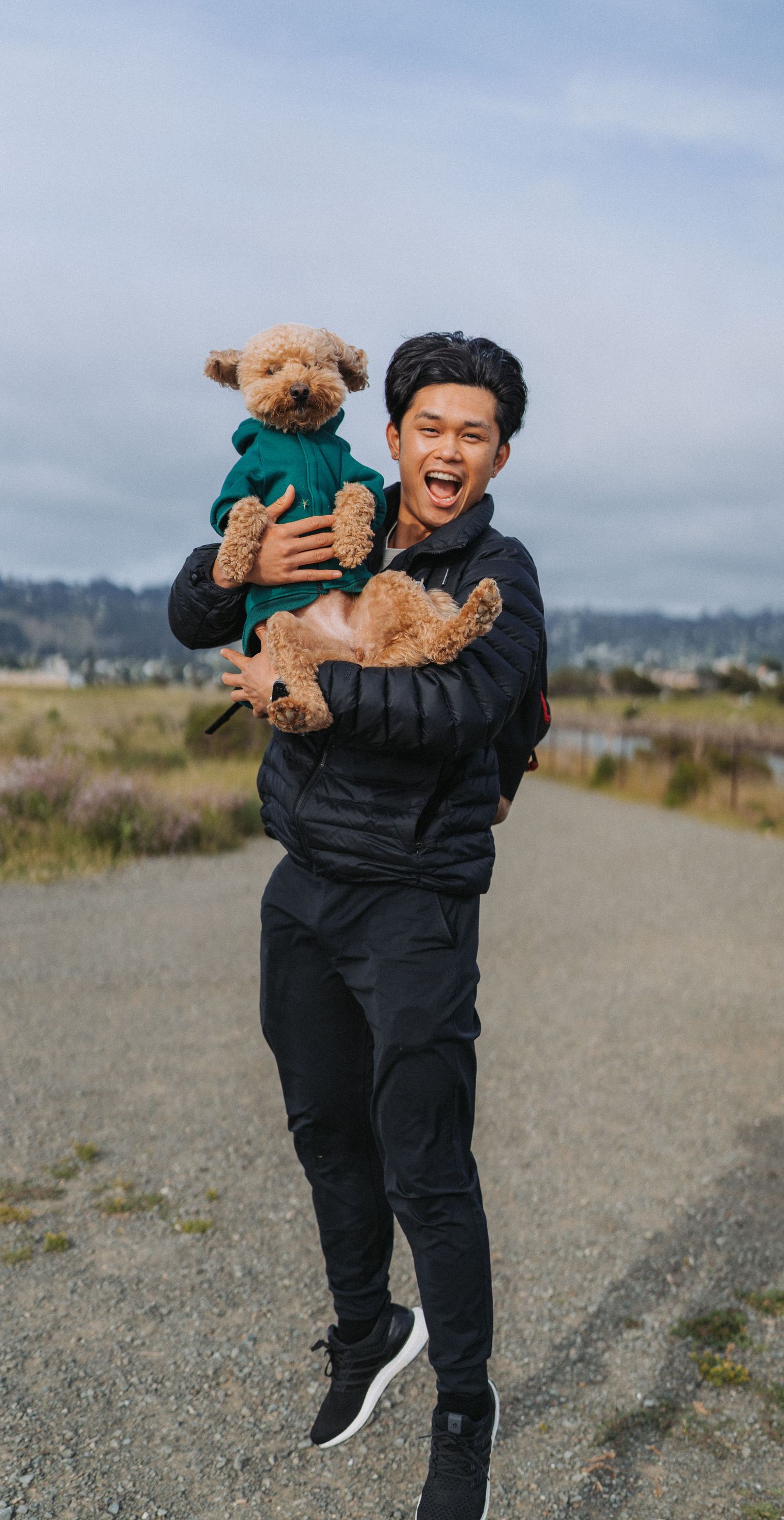 A joyful young man in black outfit holds a small brown poodle in a green hoodie outdoors on a cloudy day.