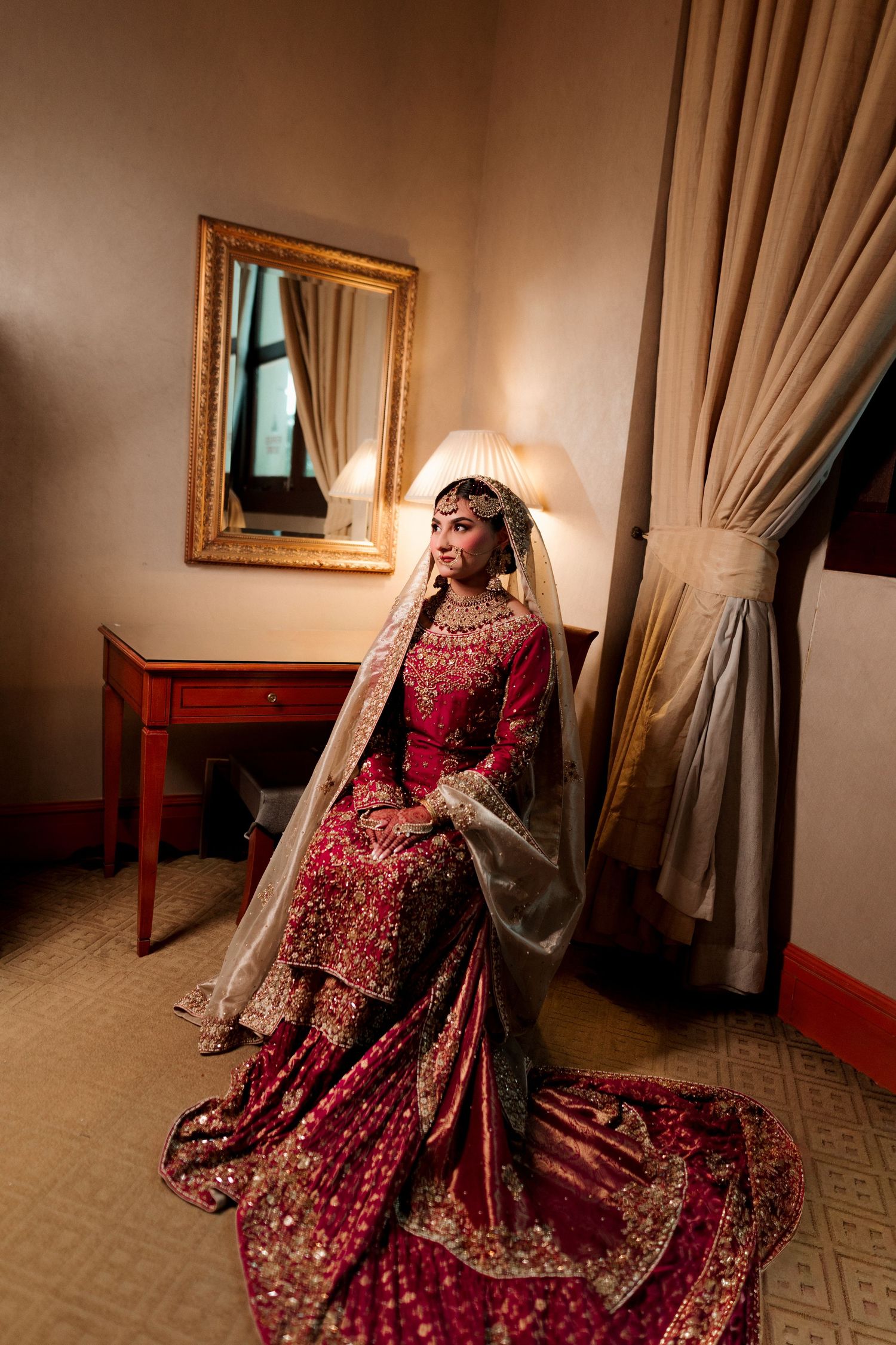 A bride in a red embroidered lehenga with gold jewelry and dupatta sits elegantly in a warmly lit bridal suite.