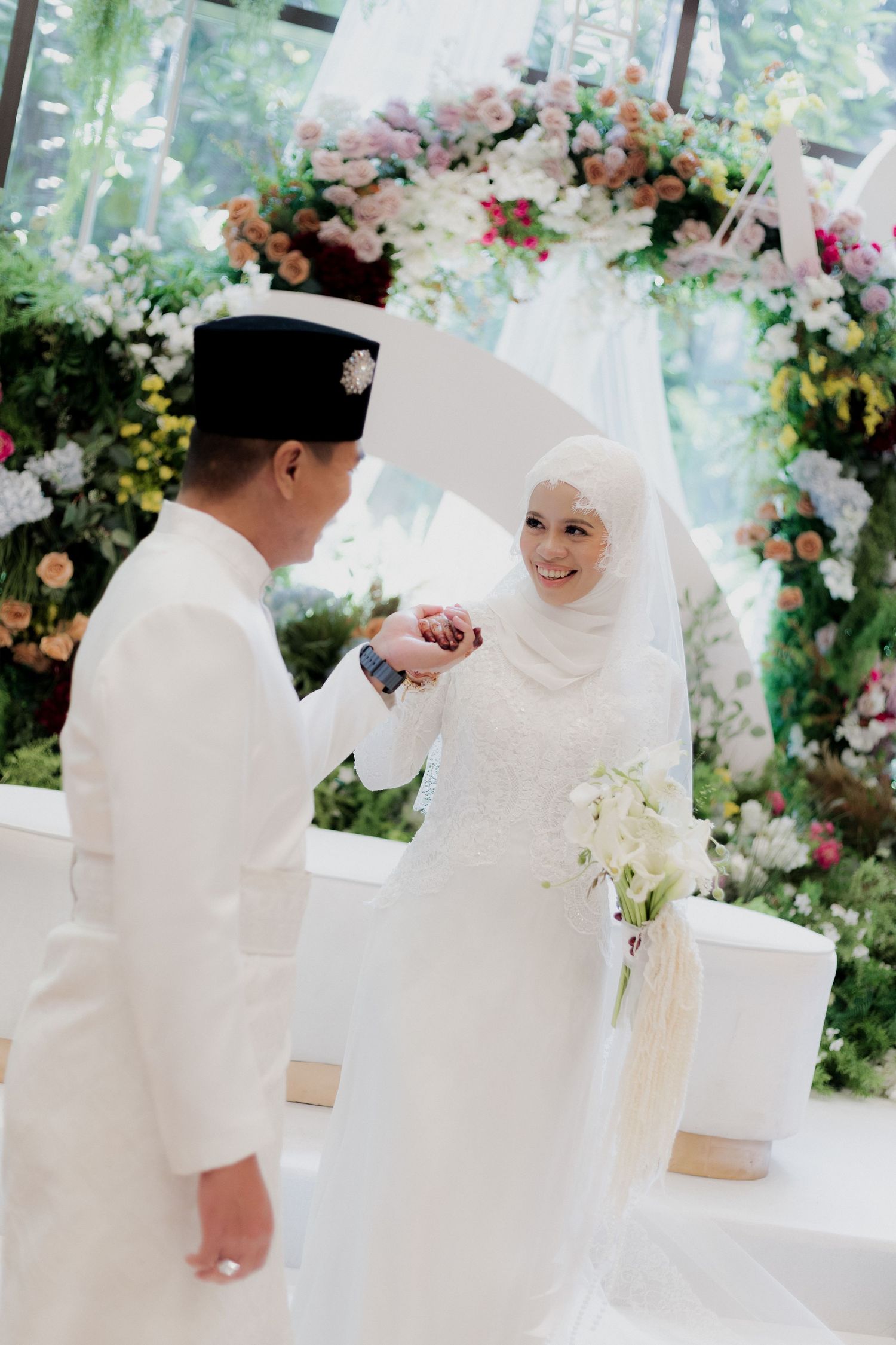 Elegant wedding ceremony with floral arch featuring white and pink blooms as couple exchanges rings in traditional attire.