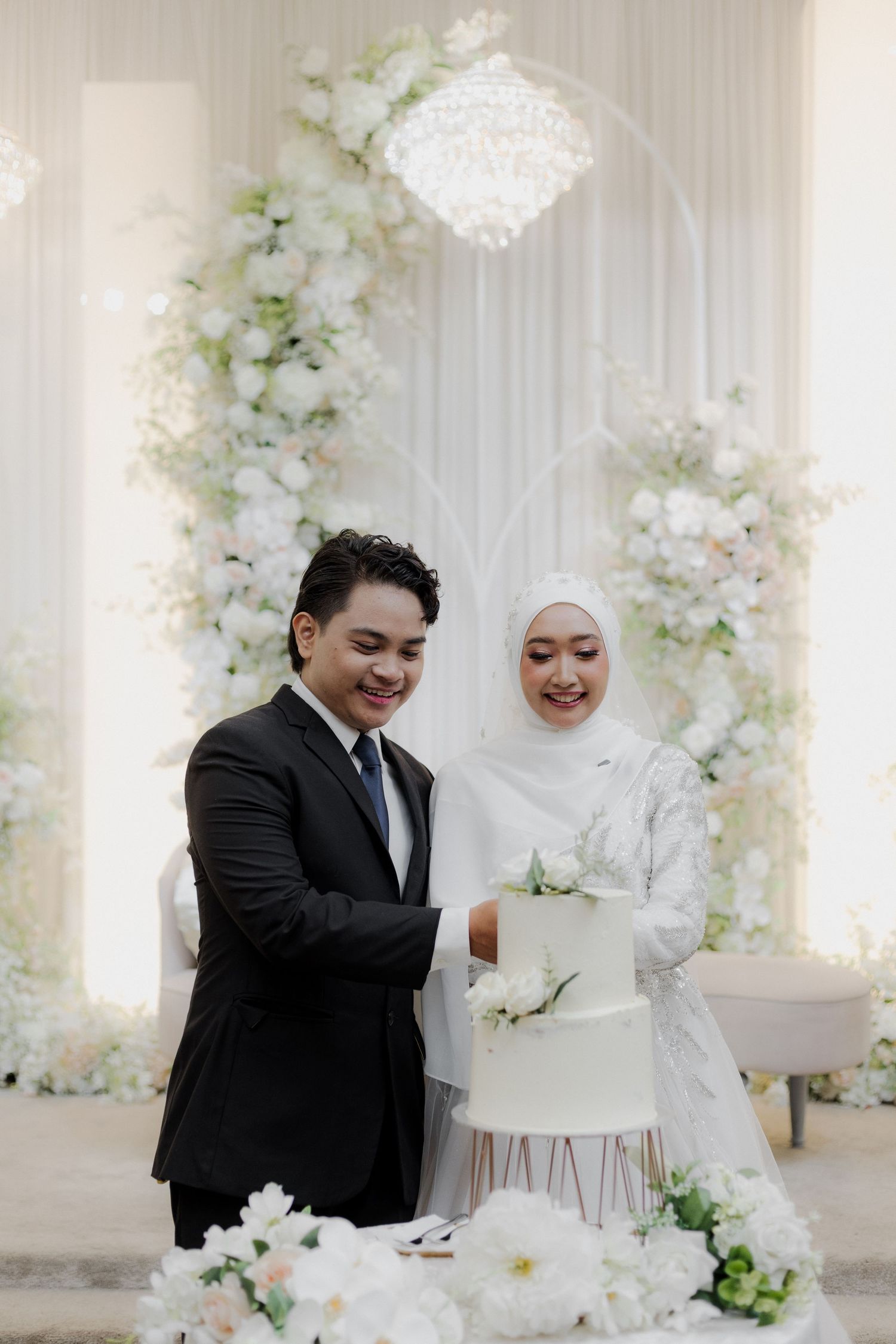 Muslim bride in hijab and groom in black suit cutting white wedding cake at elegant floral ceremony.
