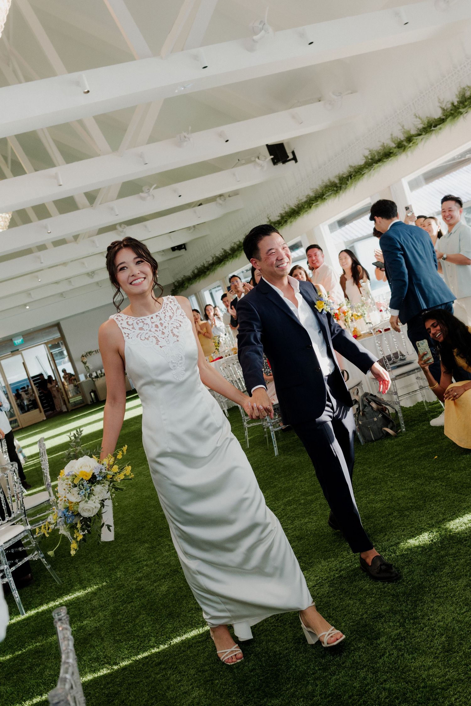 A smiling bride in a white gown and groom in a navy suit walk hand-in-hand at an elegant indoor wedding venue.