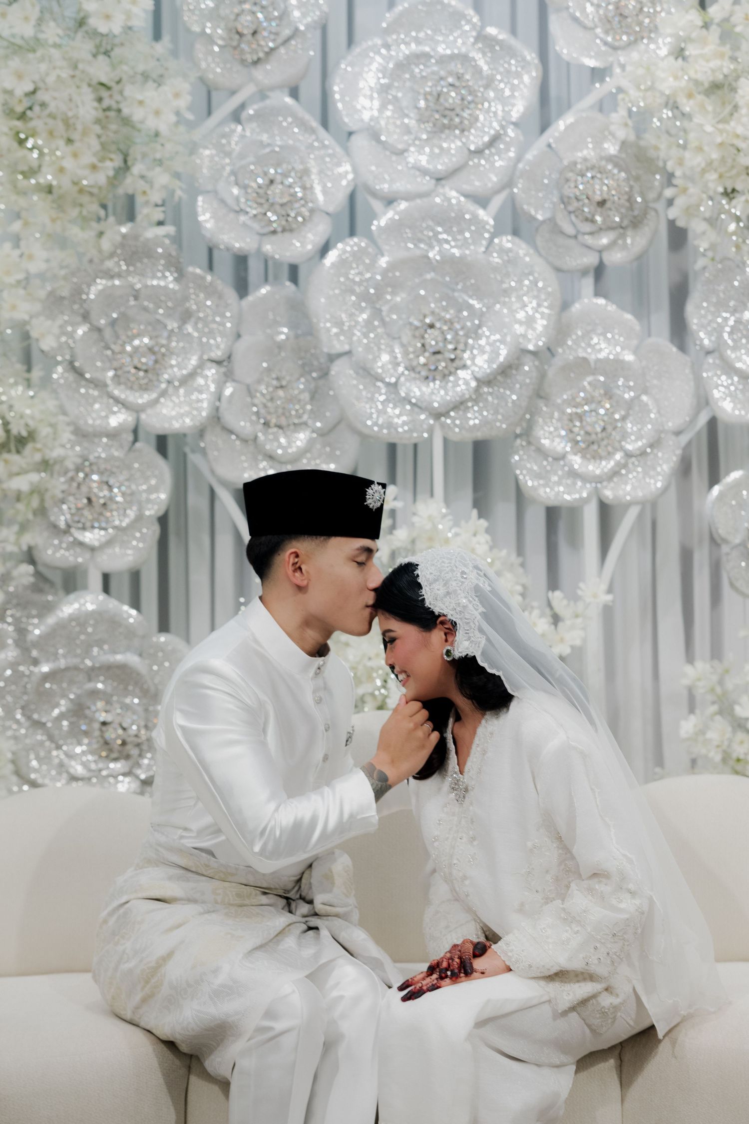 A couple in traditional white Malay wedding attire shares a romantic moment before a glittering floral backdrop.