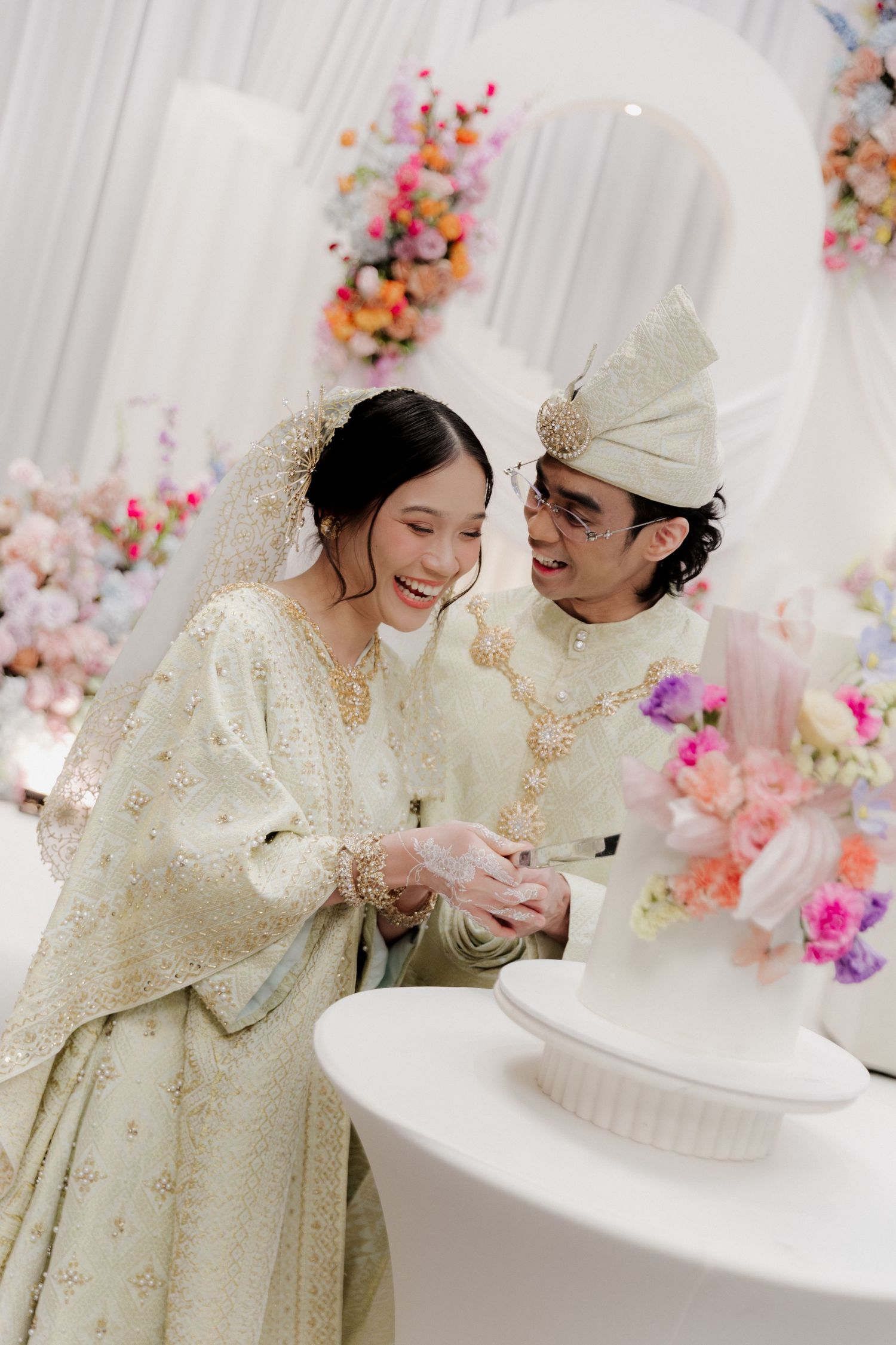 Malay couple in traditional wedding attire cutting a white cake, surrounded by colorful floral decorations.