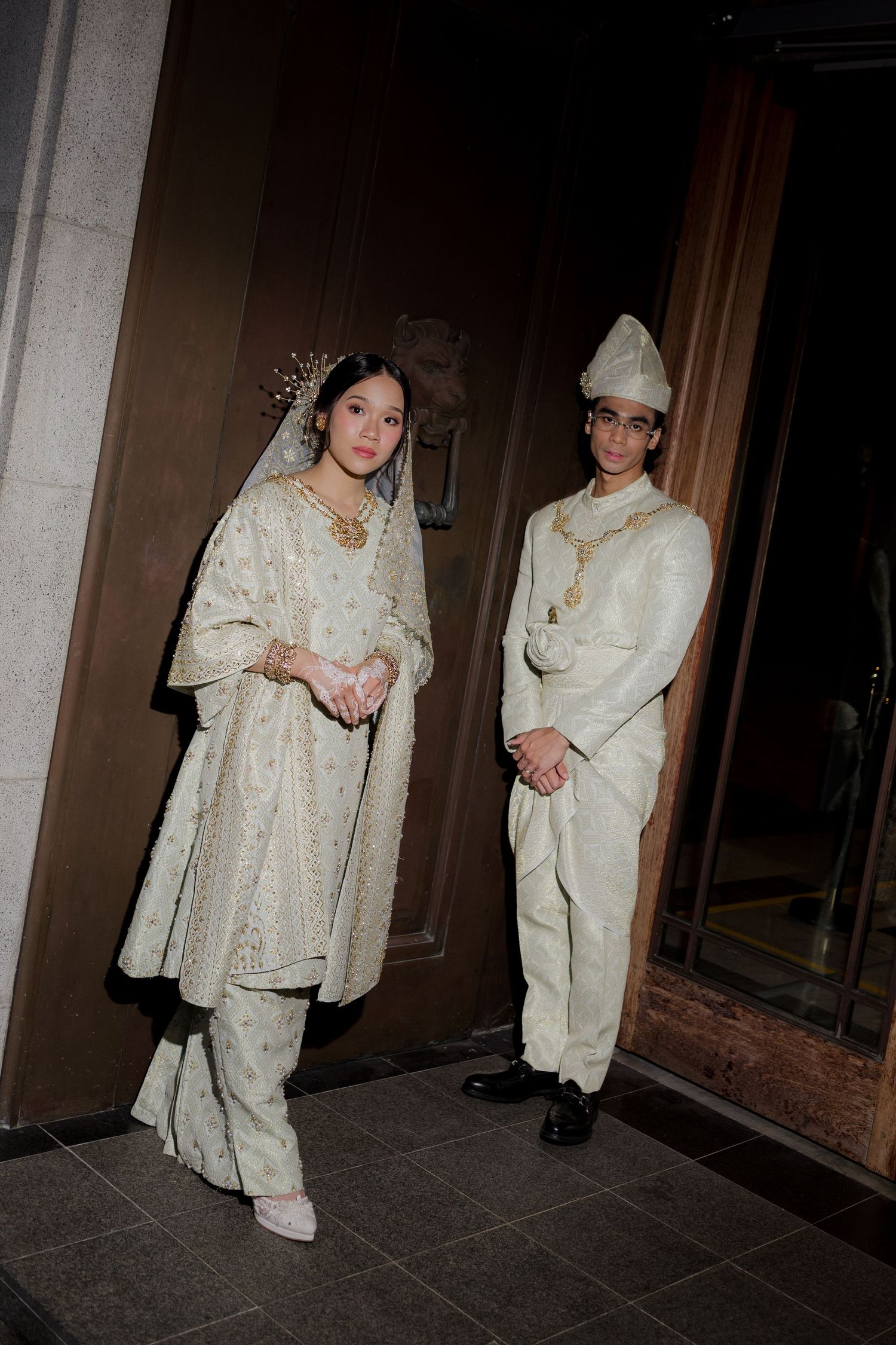 A couple in traditional Malay wedding attire, wearing matching ivory and gold outfits, posing elegantly indoors.