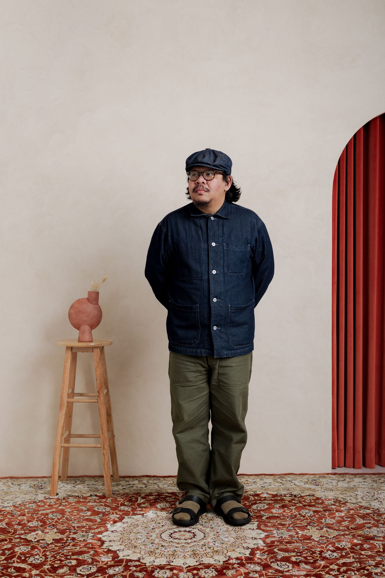 Man in navy jacket, green pants, and cap stands on a patterned rug beside a wooden stool with a vase.