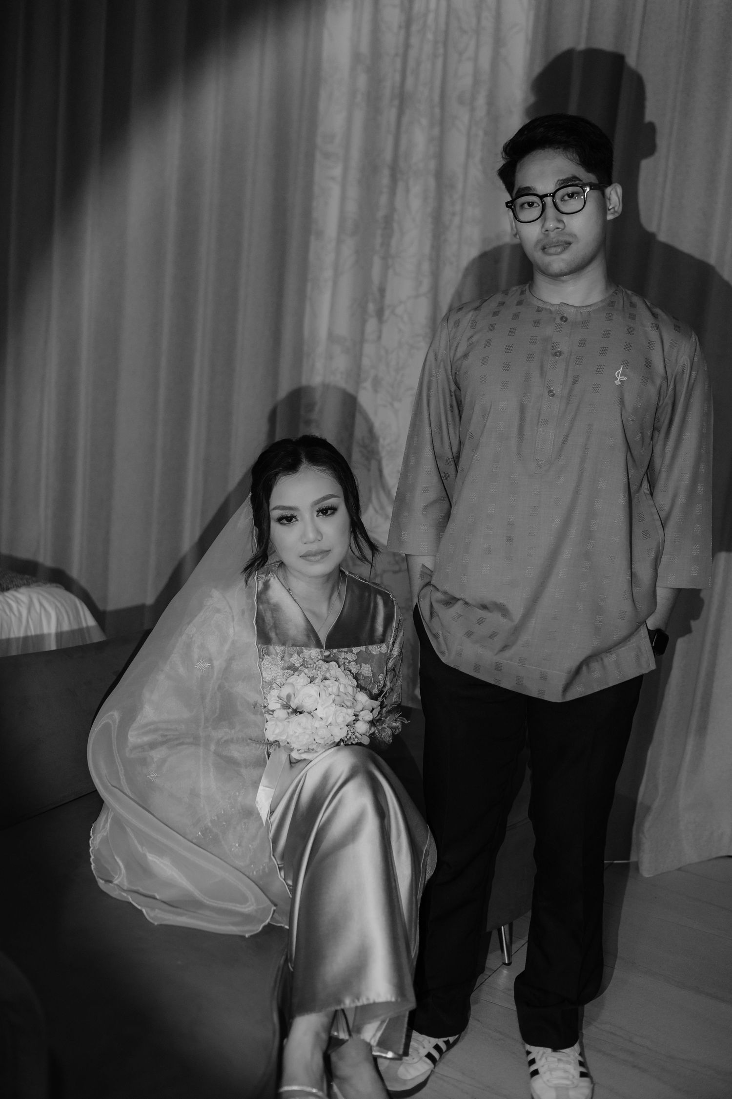 Black and white photo of a bride holding bouquet seated beside groom in traditional Malay attire indoors.