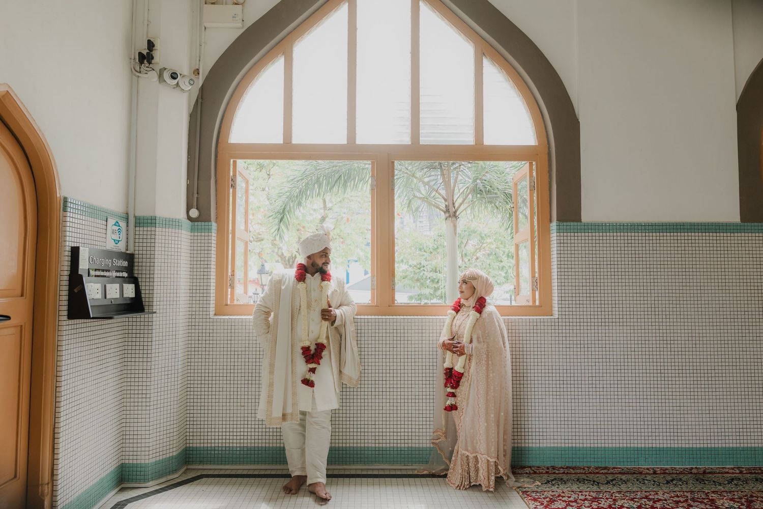 Bride and groom in traditional South Asian wedding attire standing in an elegant mosque with arched windows.