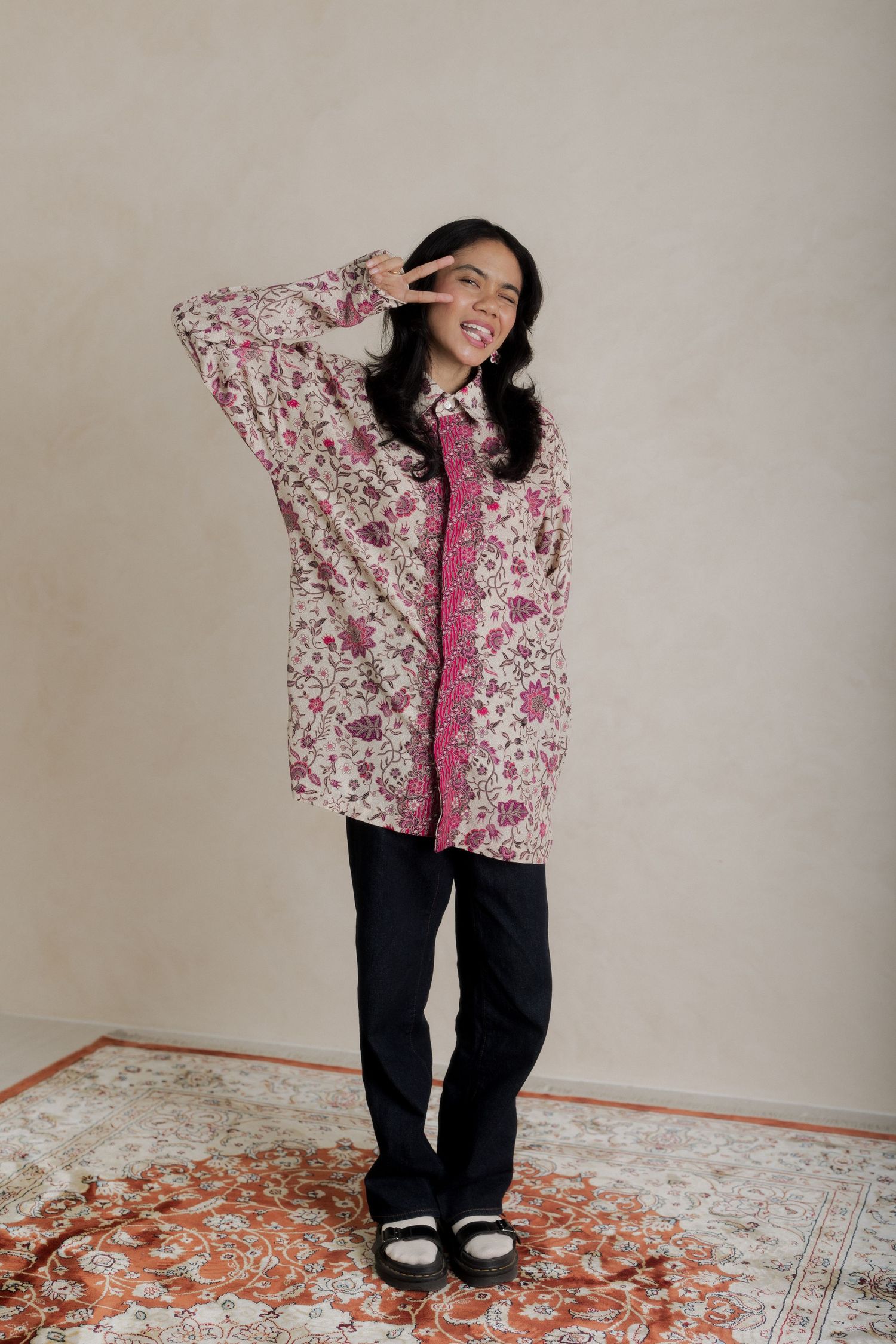 Woman posing in floral pink oversized shirt, black trousers, and platform shoes, making a peace sign.