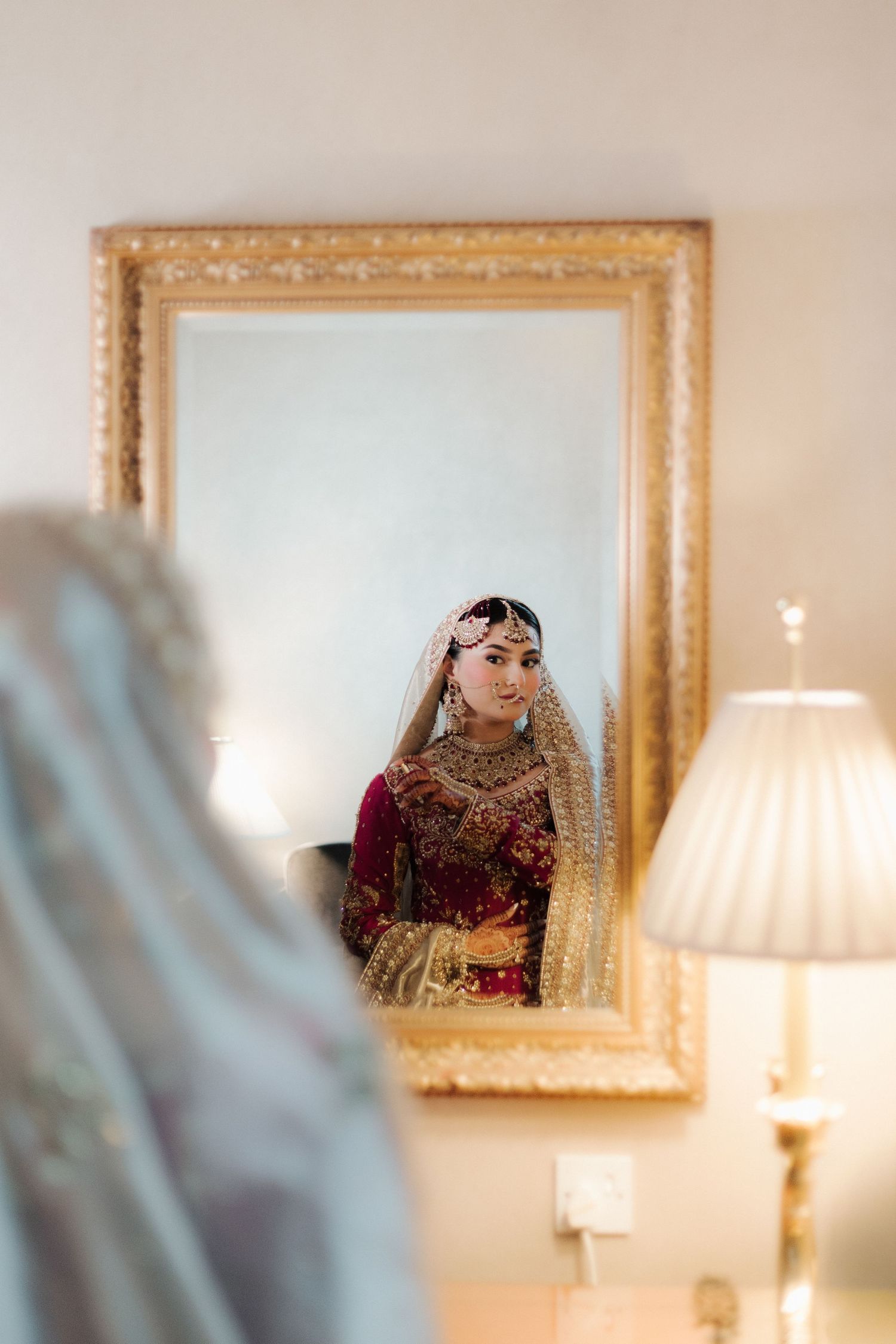 Bride in traditional South Asian bridal attire reflected in ornate gold mirror, wearing red lehenga and gold jewelry.