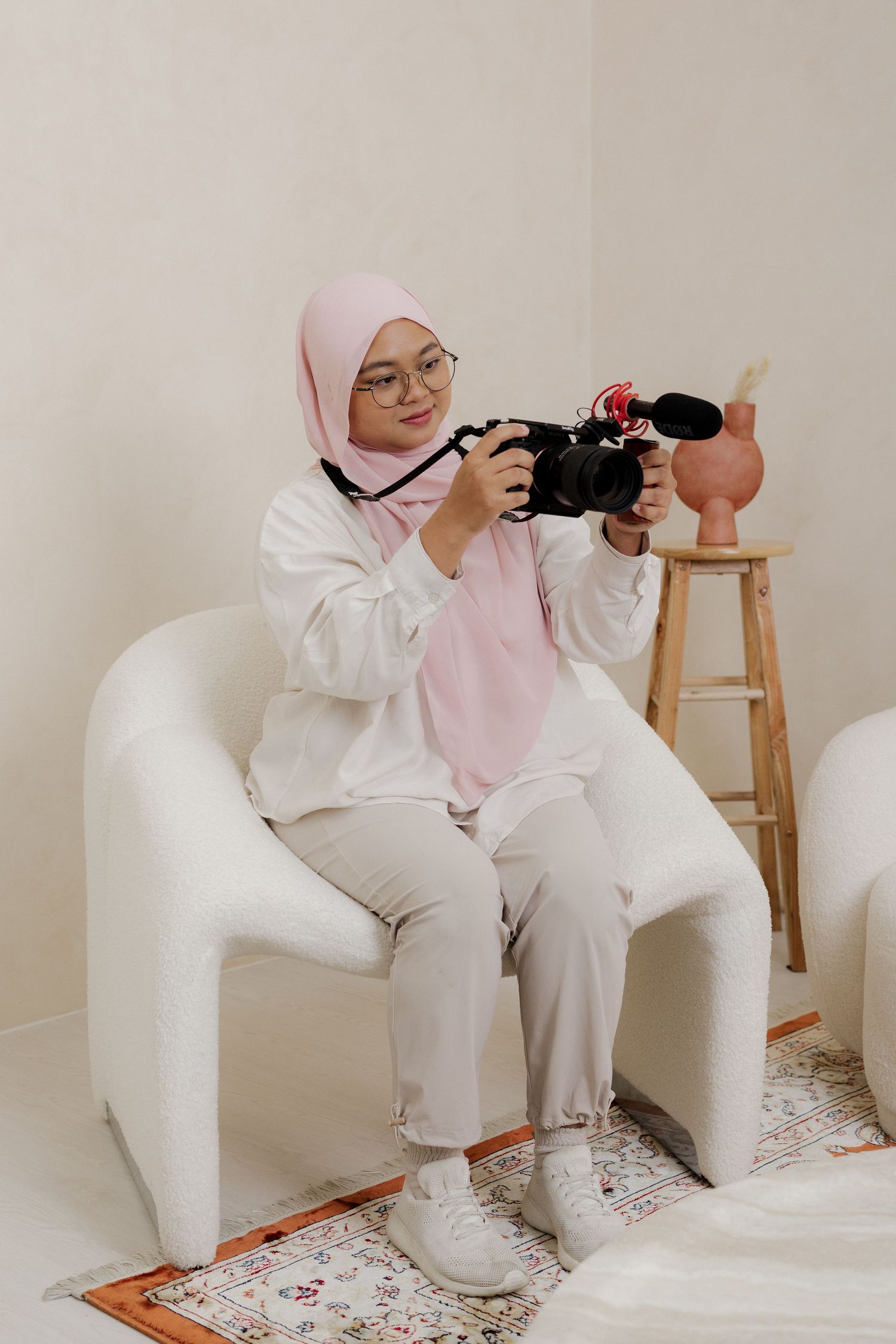 Young Muslim woman in pink hijab and glasses sits in white chair holding DSLR camera with microphone attachment.