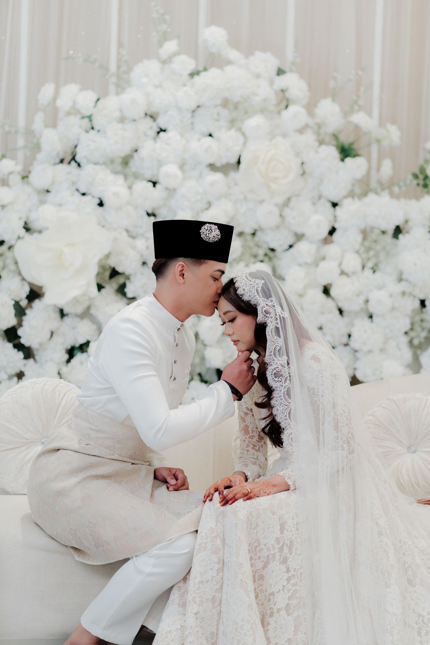 Malay couple in white traditional attire sharing a tender moment at an elegant wedding with white floral backdrop.