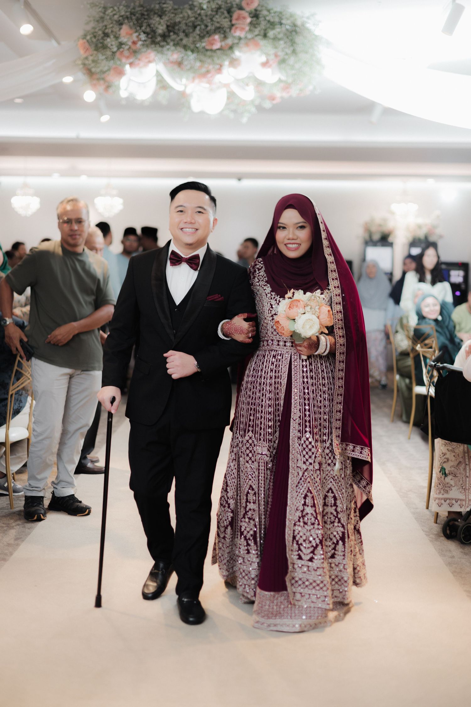 Muslim couple walks down wedding aisle, groom in black tuxedo with cane, bride in burgundy embroidered gown and hijab.