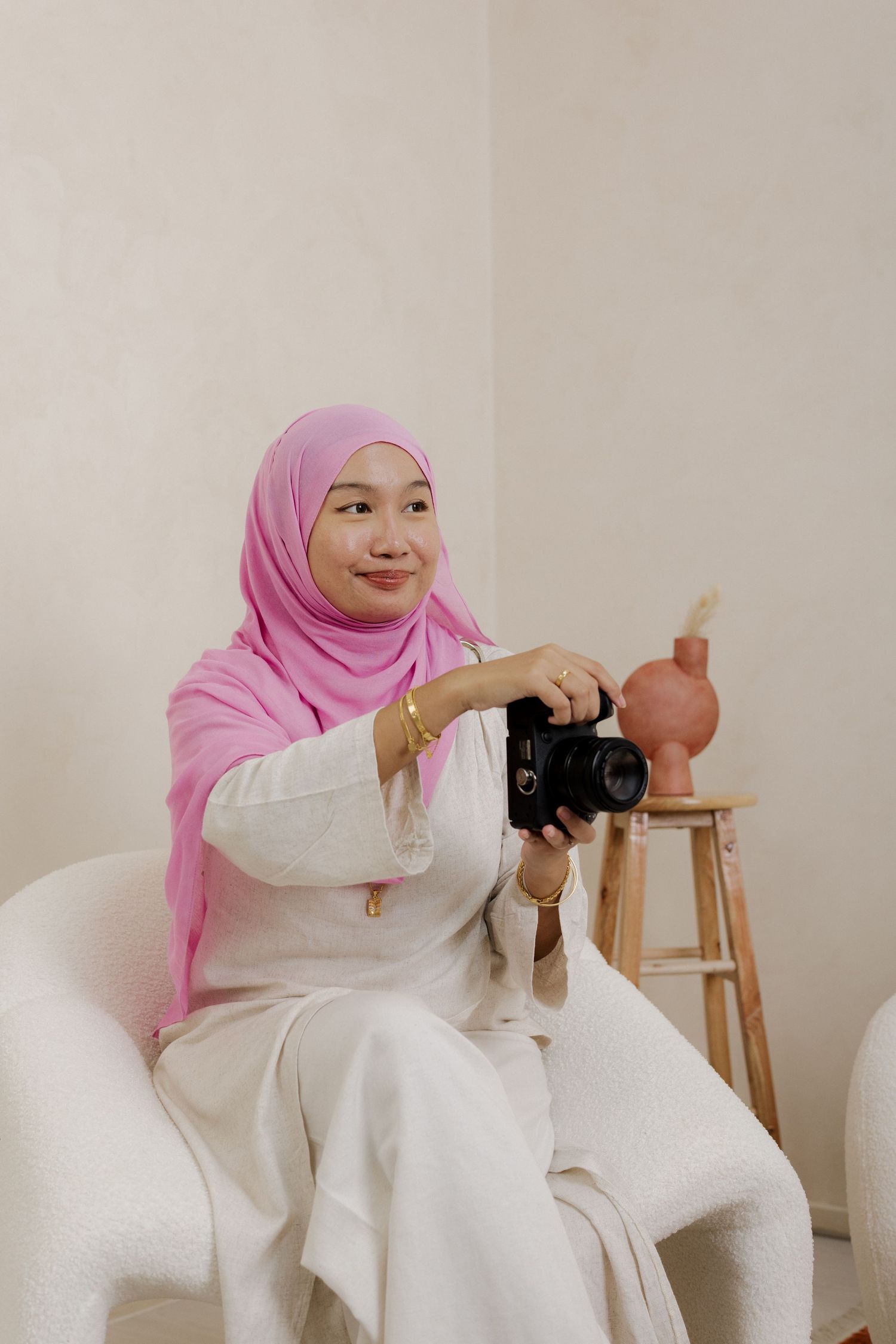 Young woman in pink hijab and white outfit sits in chair holding a DSLR camera, smiling in a bright studio.