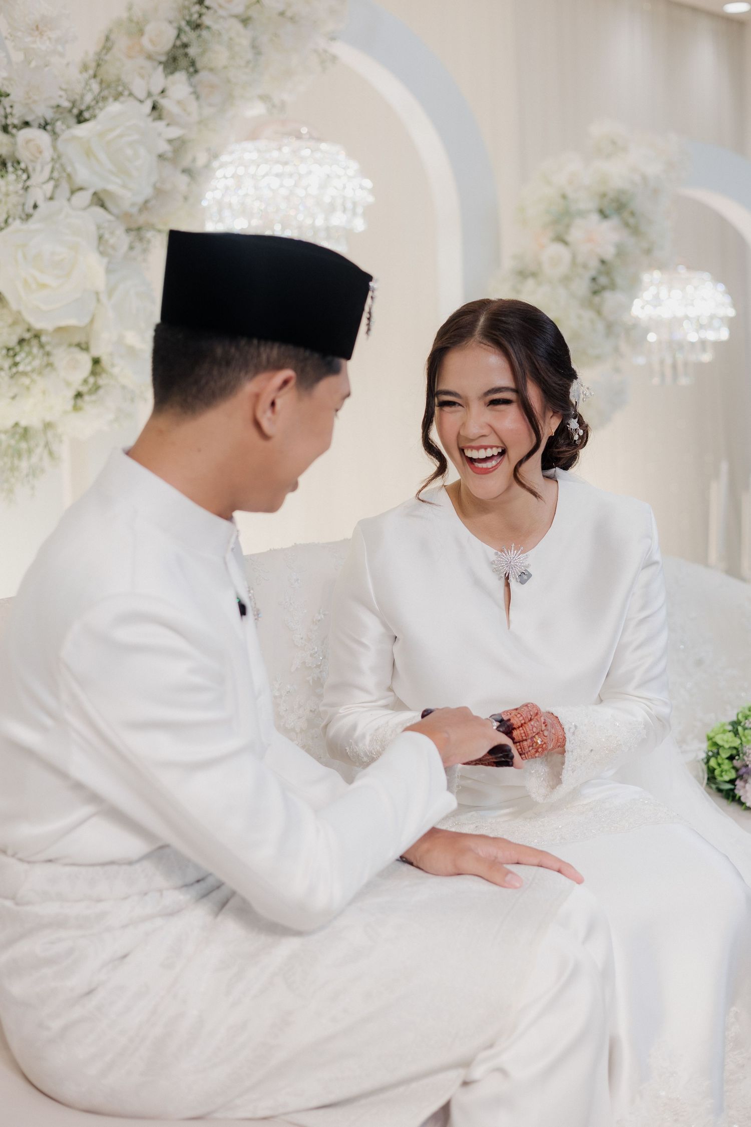 A joyful Malay couple in white traditional attire sharing a happy moment during their wedding ceremony.