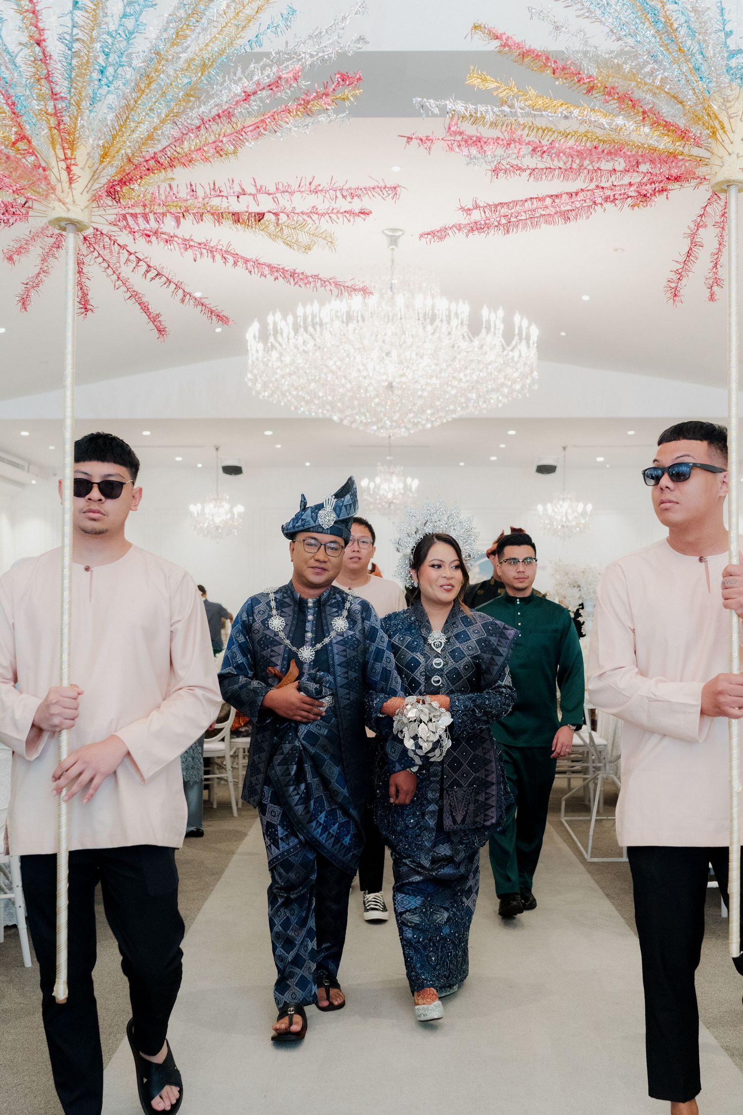 Malay wedding couple in traditional blue batik attire walks down aisle flanked by groomsmen holding decorative fans.