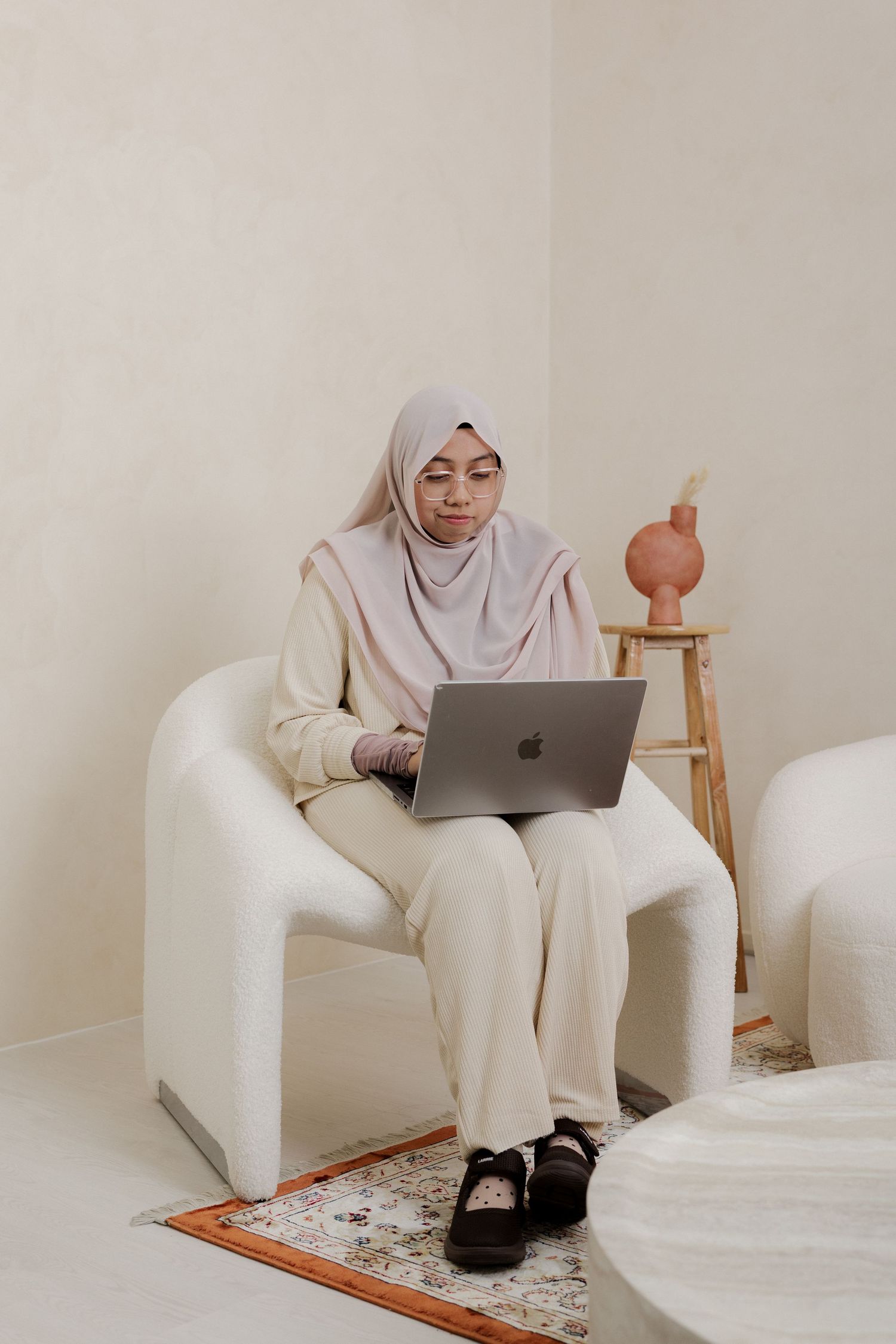 A Muslim woman in hijab sits in a white armchair working on a MacBook laptop in a minimalist beige room.