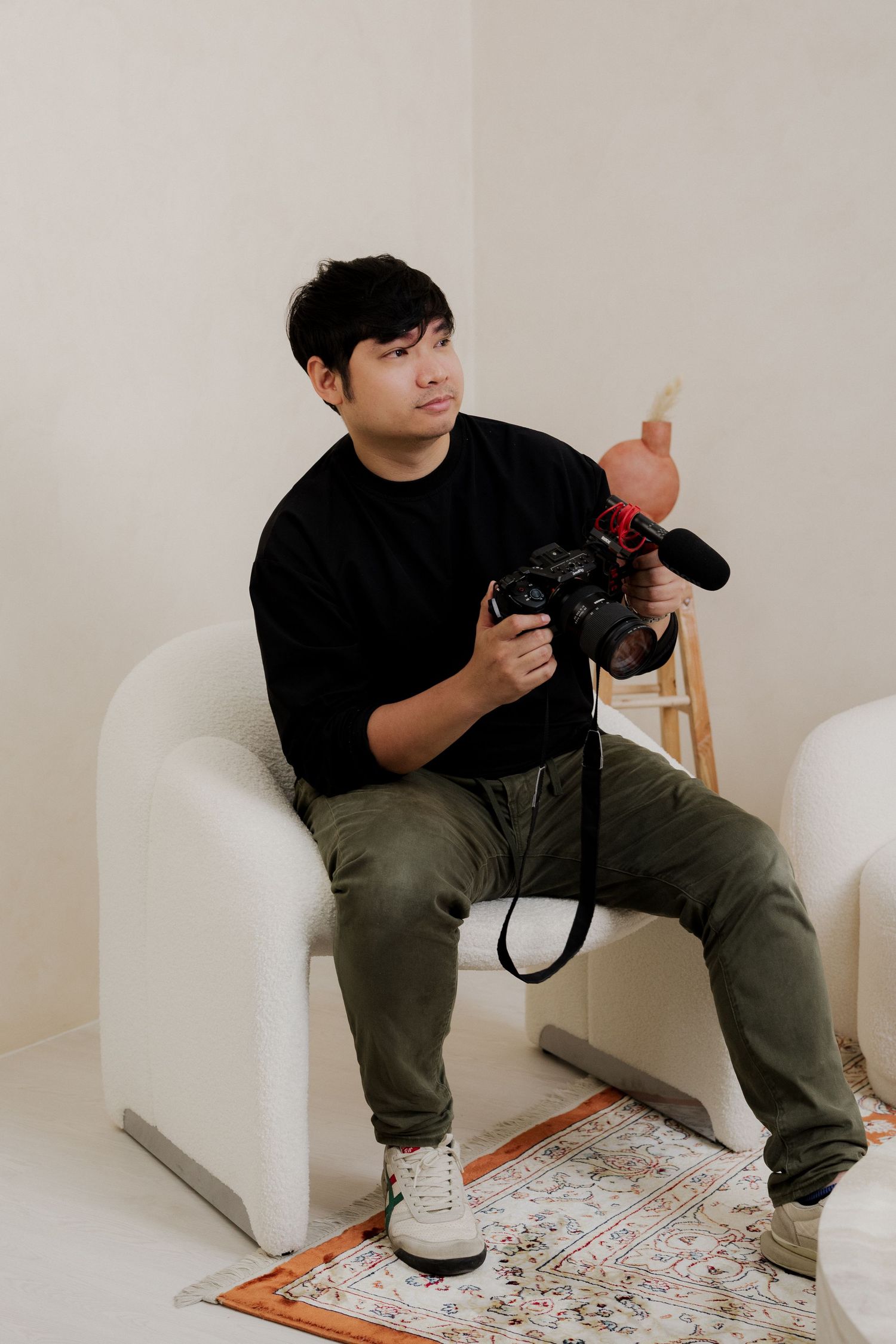 Young Asian man in black shirt and olive pants sits in white chair holding a professional camera with microphone attachment.