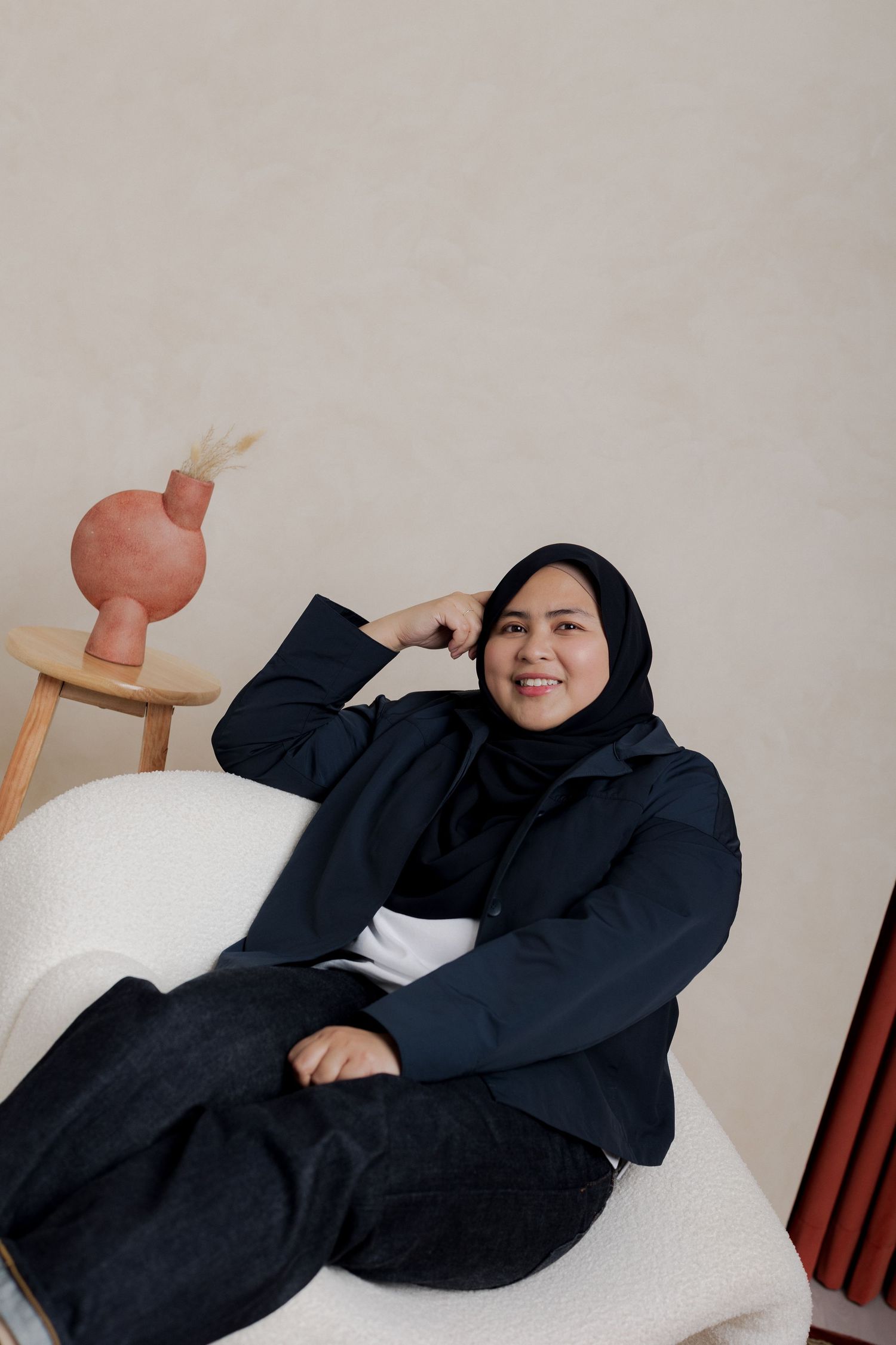 A smiling woman in a black hijab and jacket sits relaxed in a white chair beside a terracotta vase on a wooden stool.