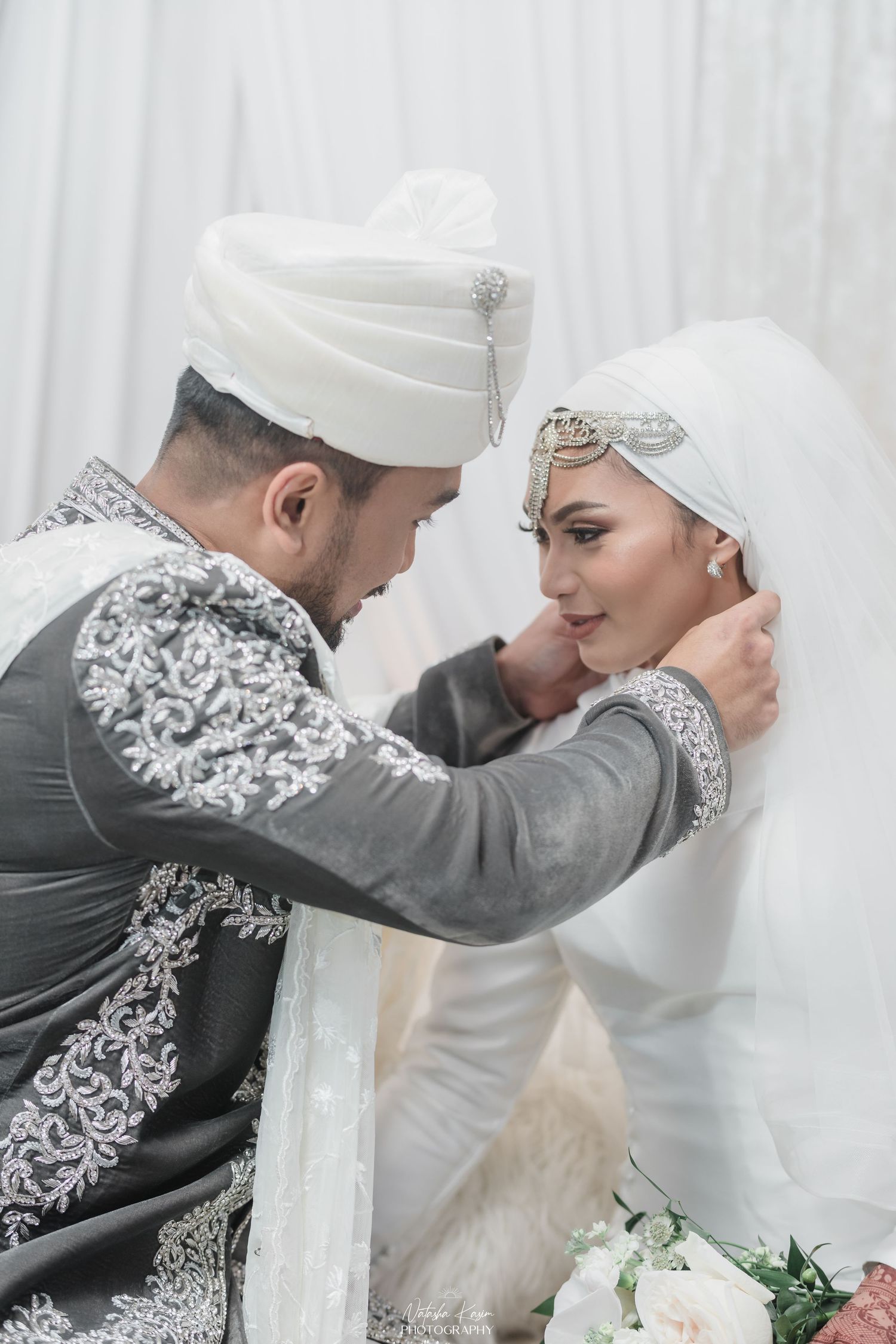 Muslim bride and groom in traditional white and gray wedding attire share an intimate moment during their ceremony.