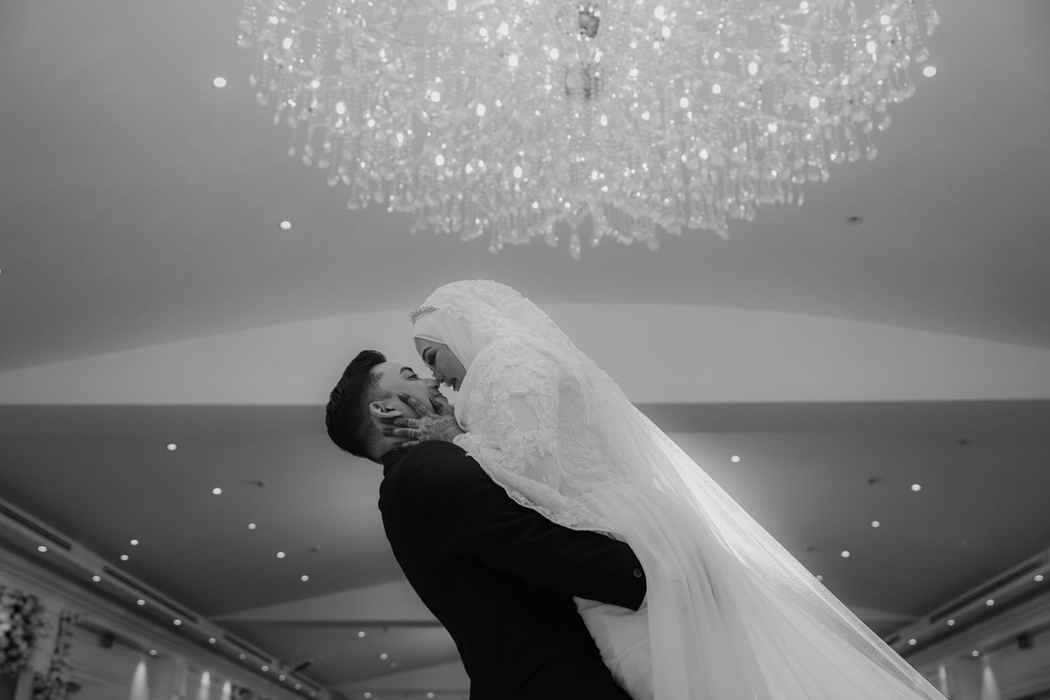 Black and white wedding photo of groom lifting bride under crystal chandelier.