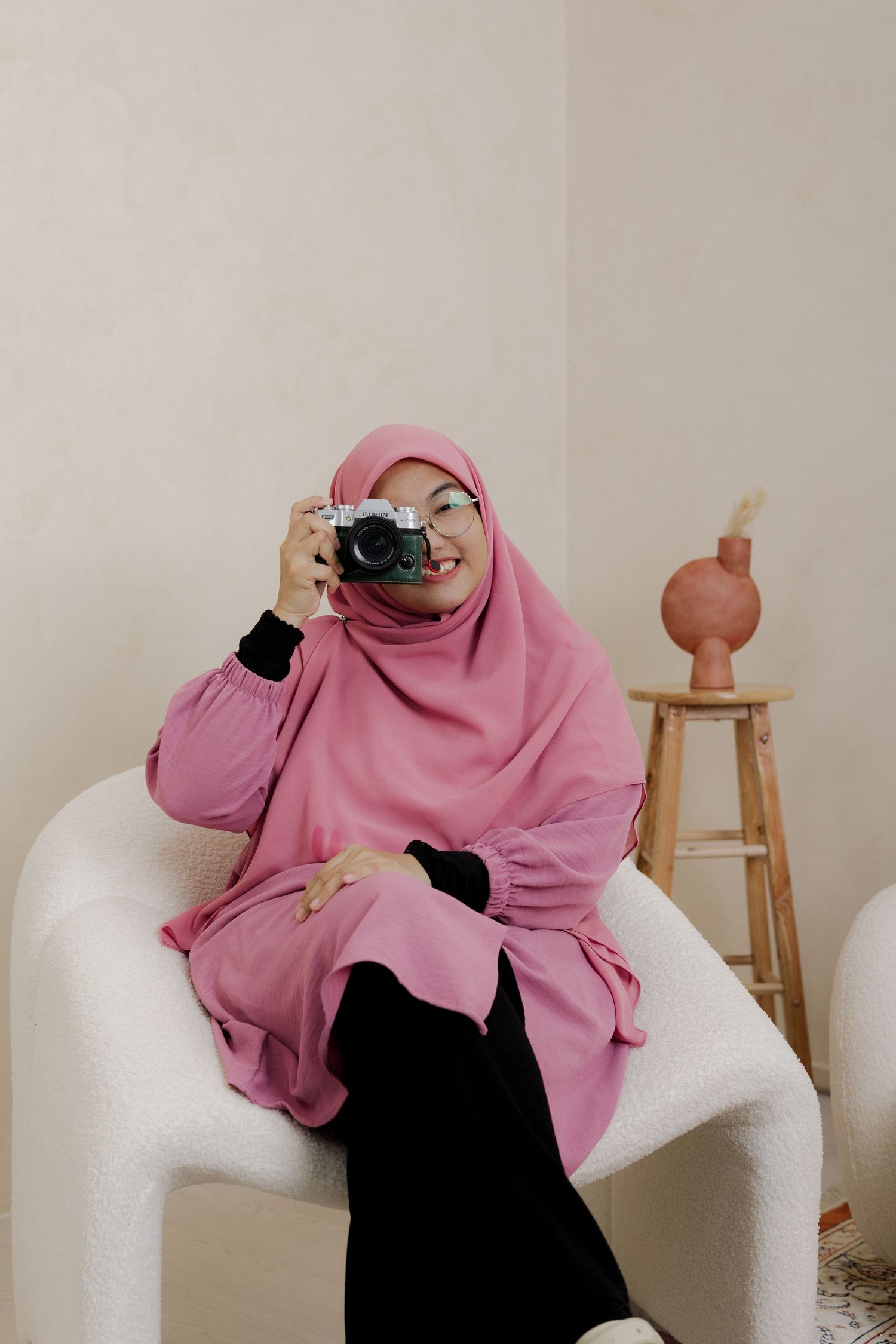 A woman in a pink hijab sits in a white chair, holding a camera up to take a photo in a minimalist room.