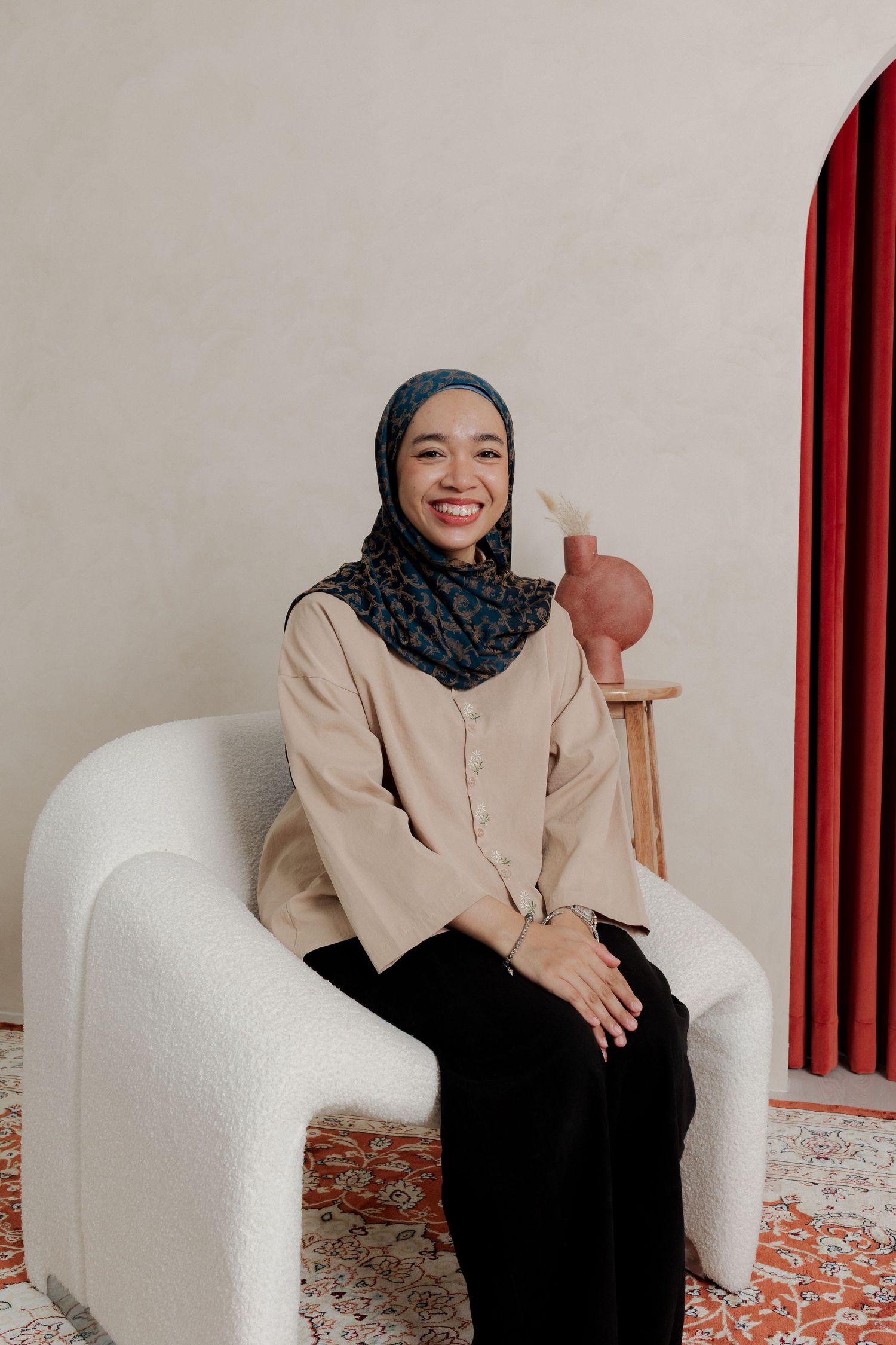 Smiling woman in hijab and beige shirt seated in white chair with decorative vase and red curtain backdrop.