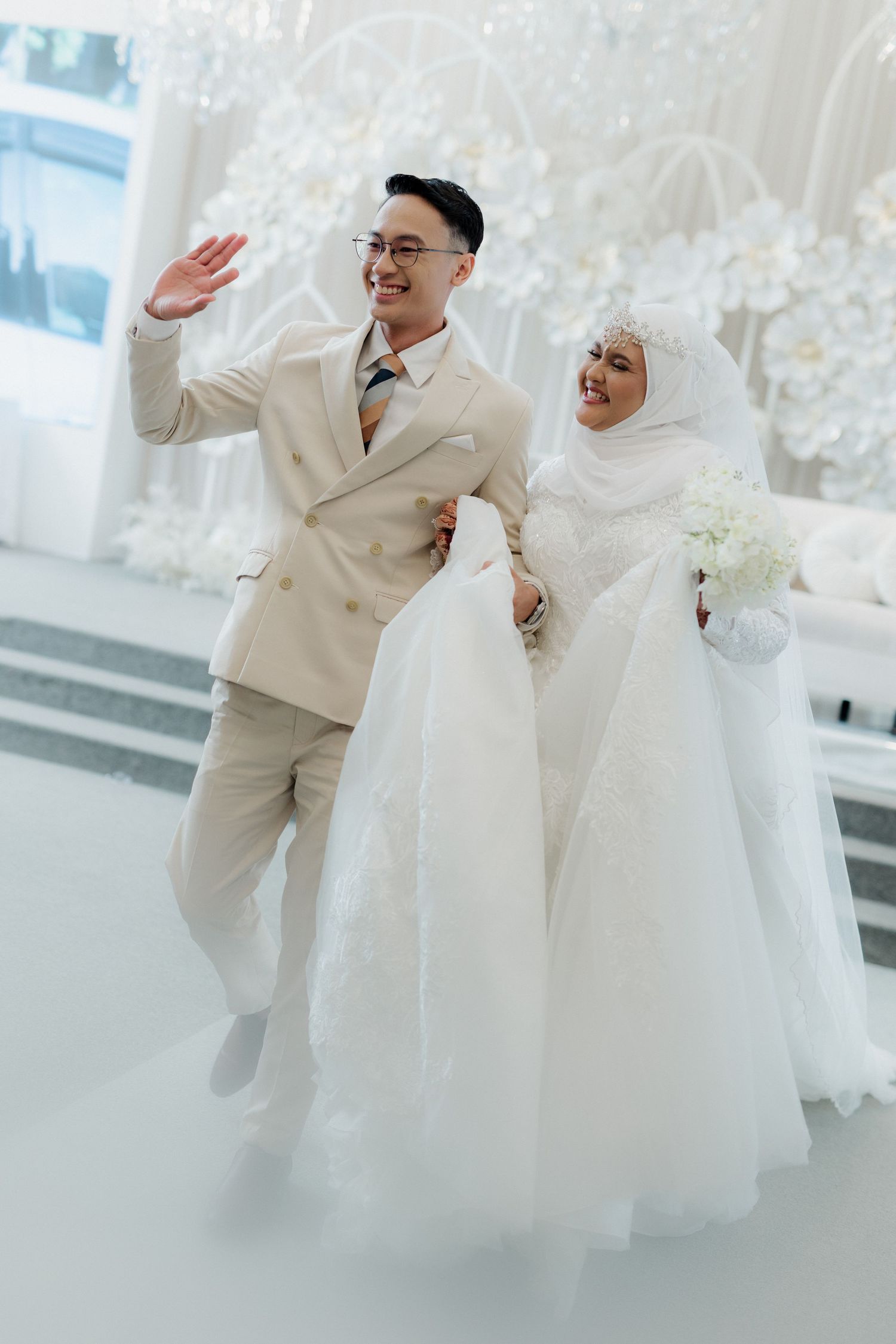 A joyful newlywed couple in white wedding attire walks together, groom waving and bride holding a bouquet at an elegant venue.