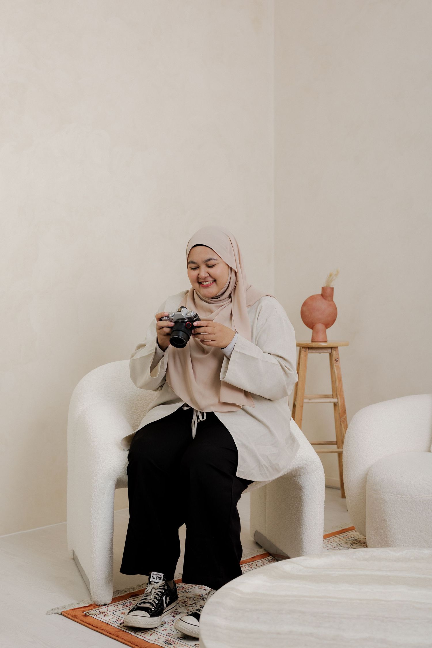 A woman in a hijab sits on a white chair, smiling while reviewing photos on a camera in a minimalist room.