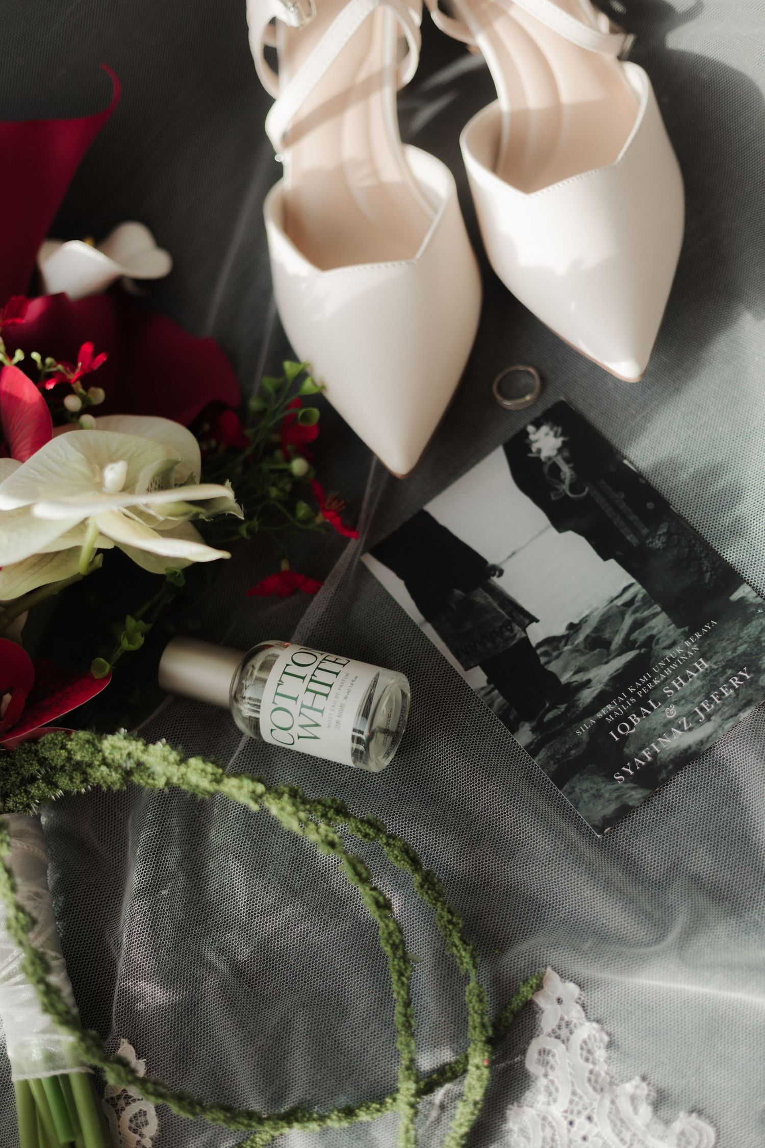 Elegant bridal flat lay with white heels, red floral bouquet, Cotton White perfume, green wreath, and a book on gray fabric.