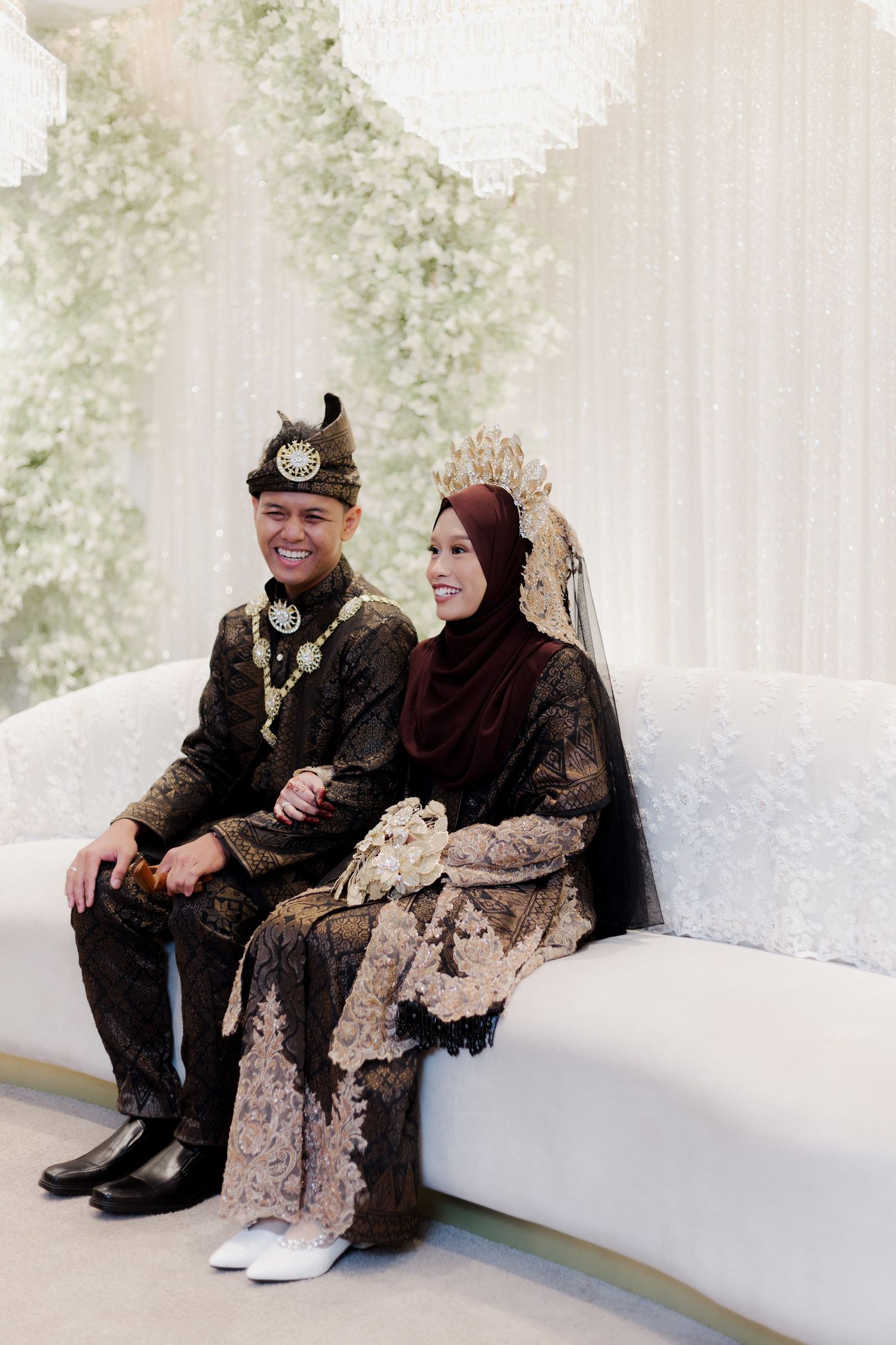 Couple in traditional Malay wedding attire seated on white sofa, groom in dark songket with tengkolok, bride in lace kebaya.