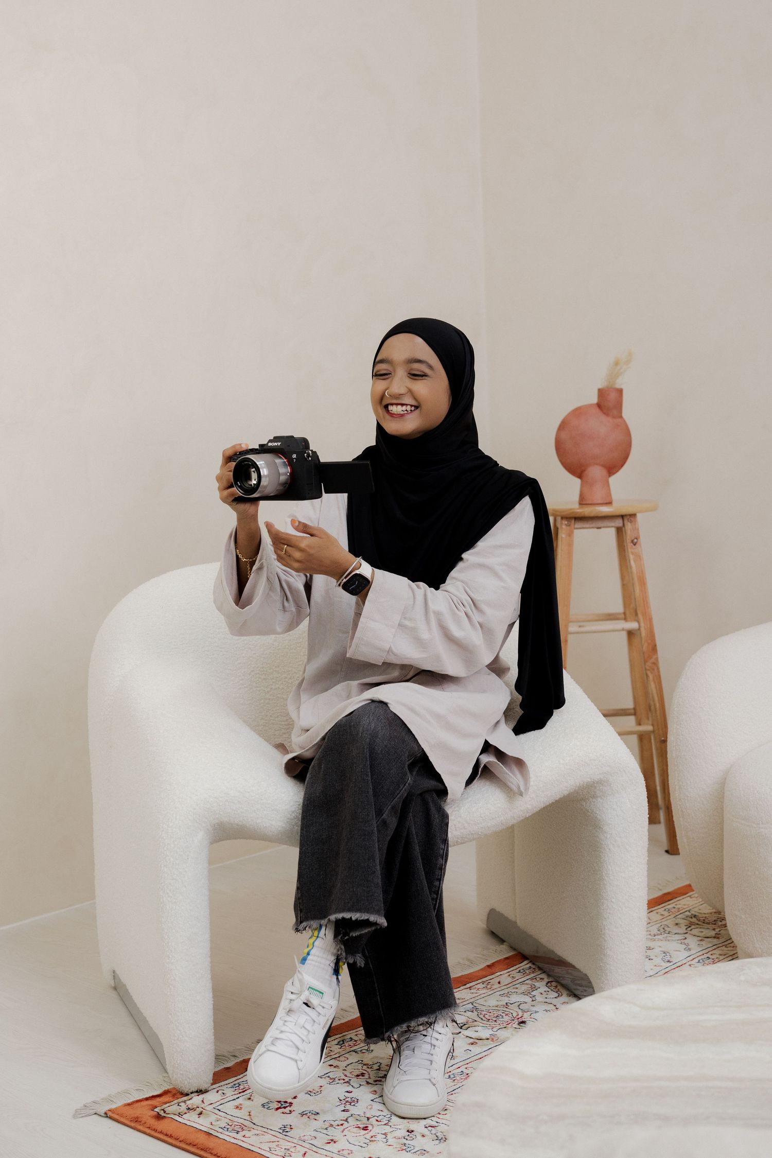 Smiling woman in black hijab sitting in white chair holding a camera in a minimalist beige studio setting.