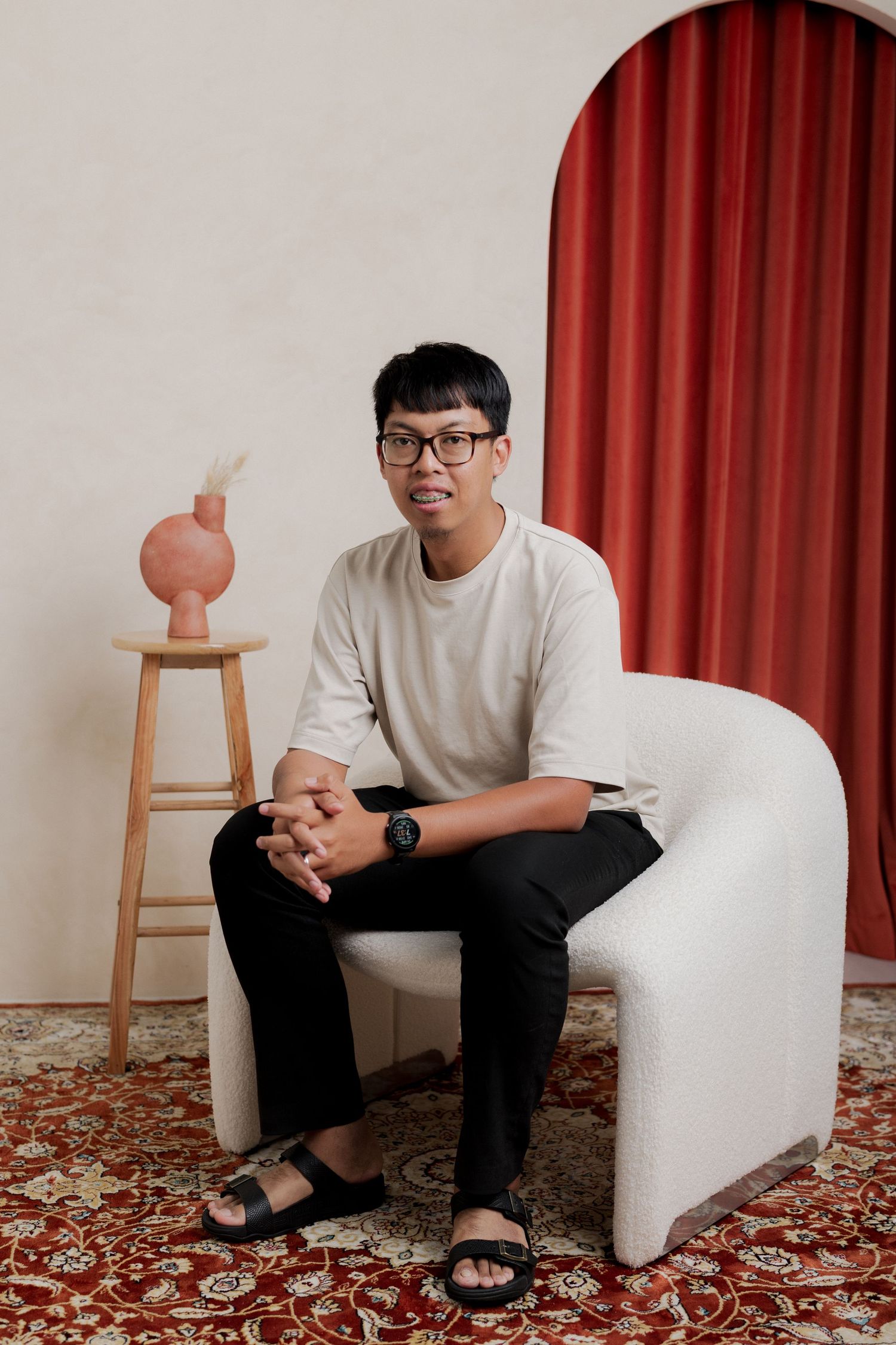 Young man in glasses and white shirt sits in a white chair beside a wooden stool with pink vase, red curtain backdrop.
